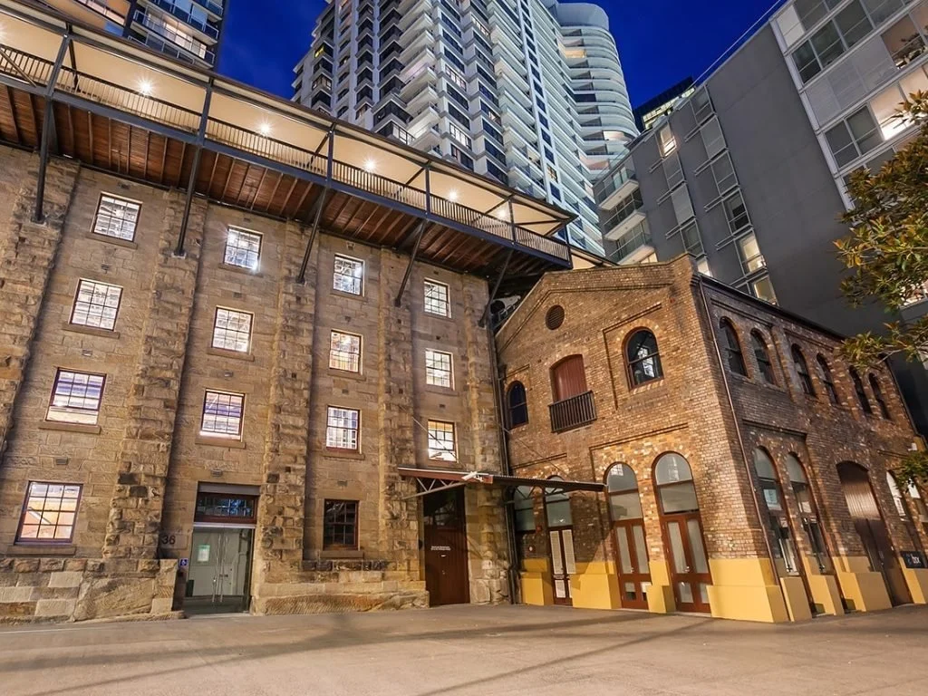 A nighttime street view showing a historic brick building with arched windows and a modern high-rise building behind it, illuminated and set against a dark blue sky.