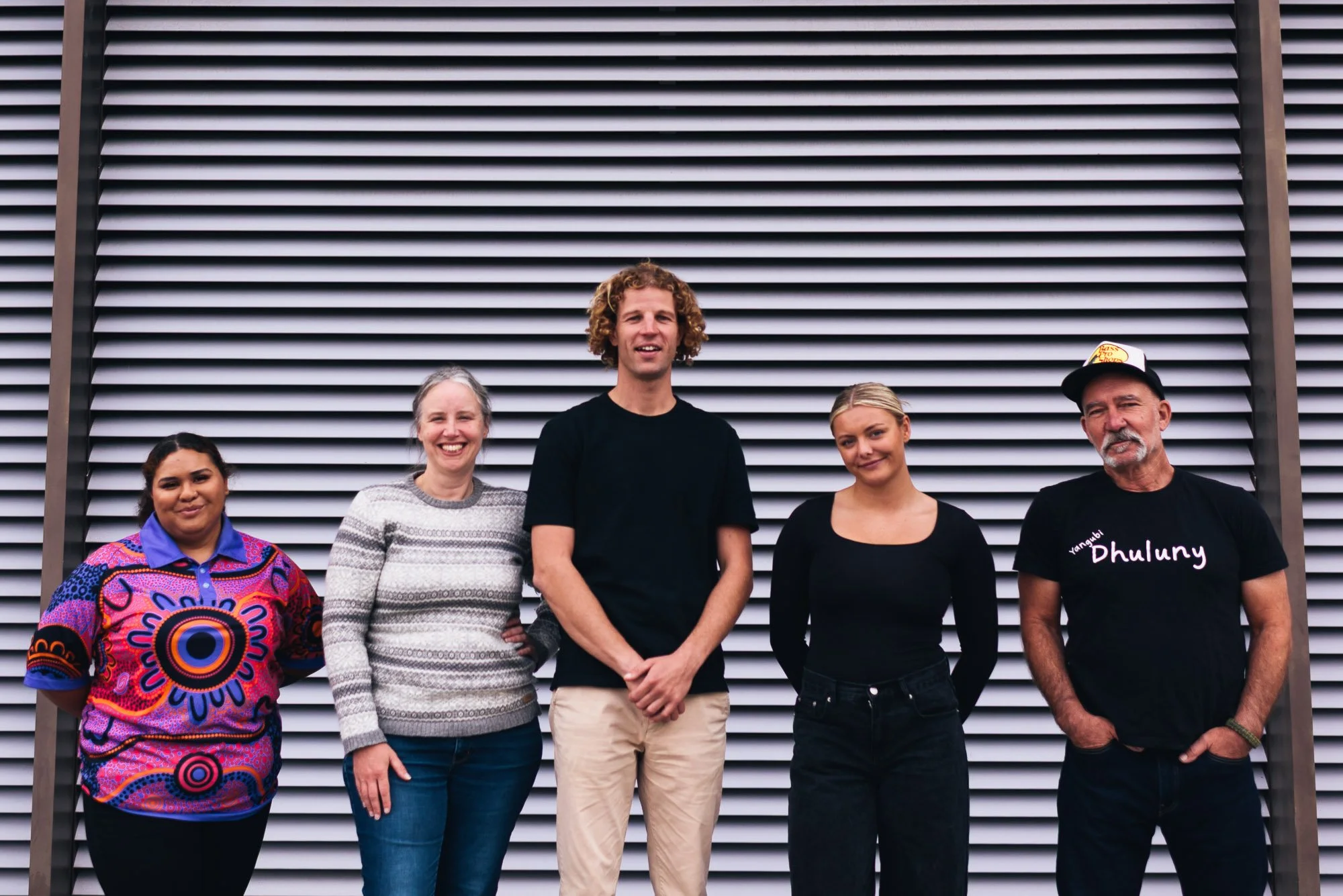 Group of five diverse people standing in front of an industrial-style wall with horizontal metal slats, smiling and posing for the photo.
