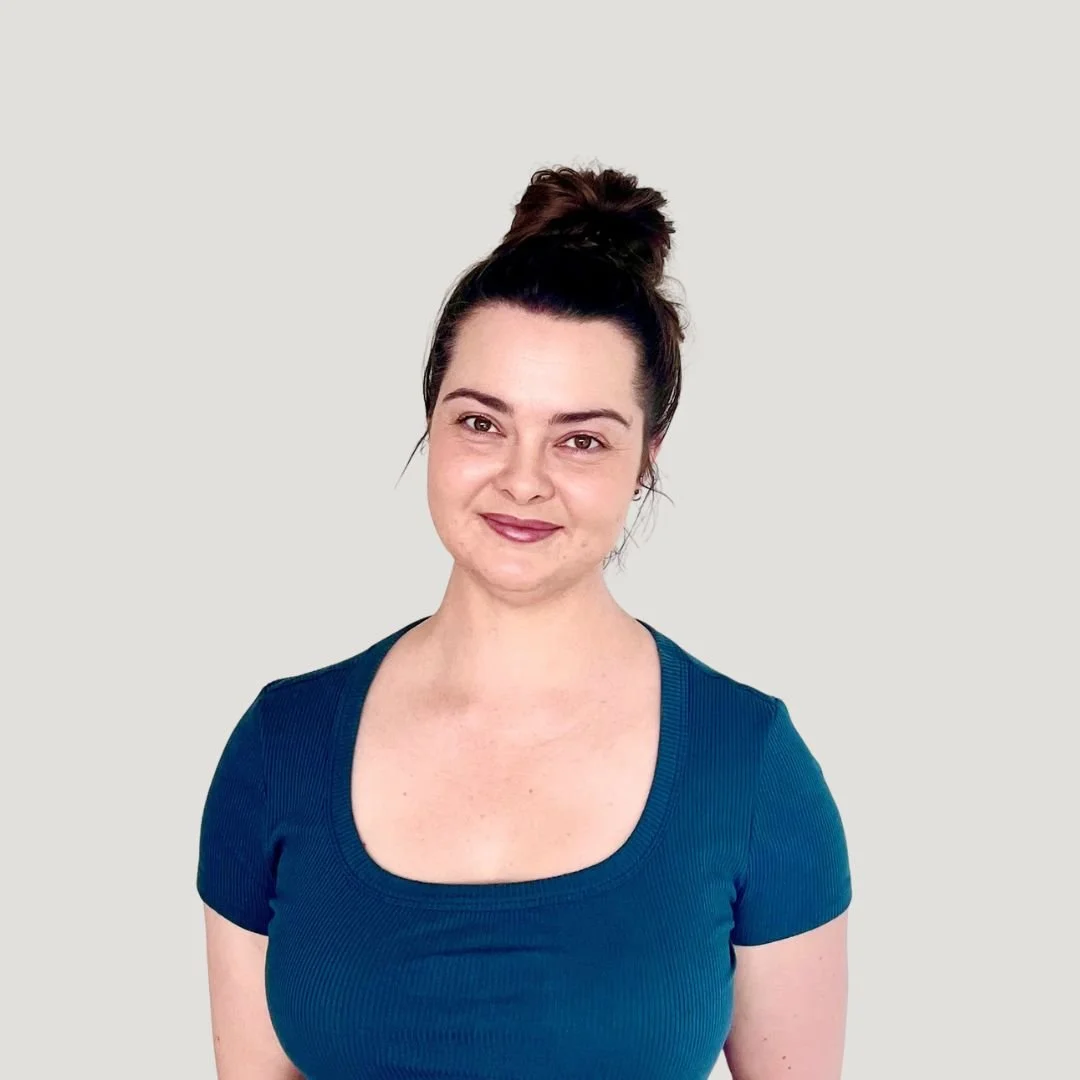 A woman with dark hair in a bun, wearing a blue top, smiling gently, standing against a plain light background.