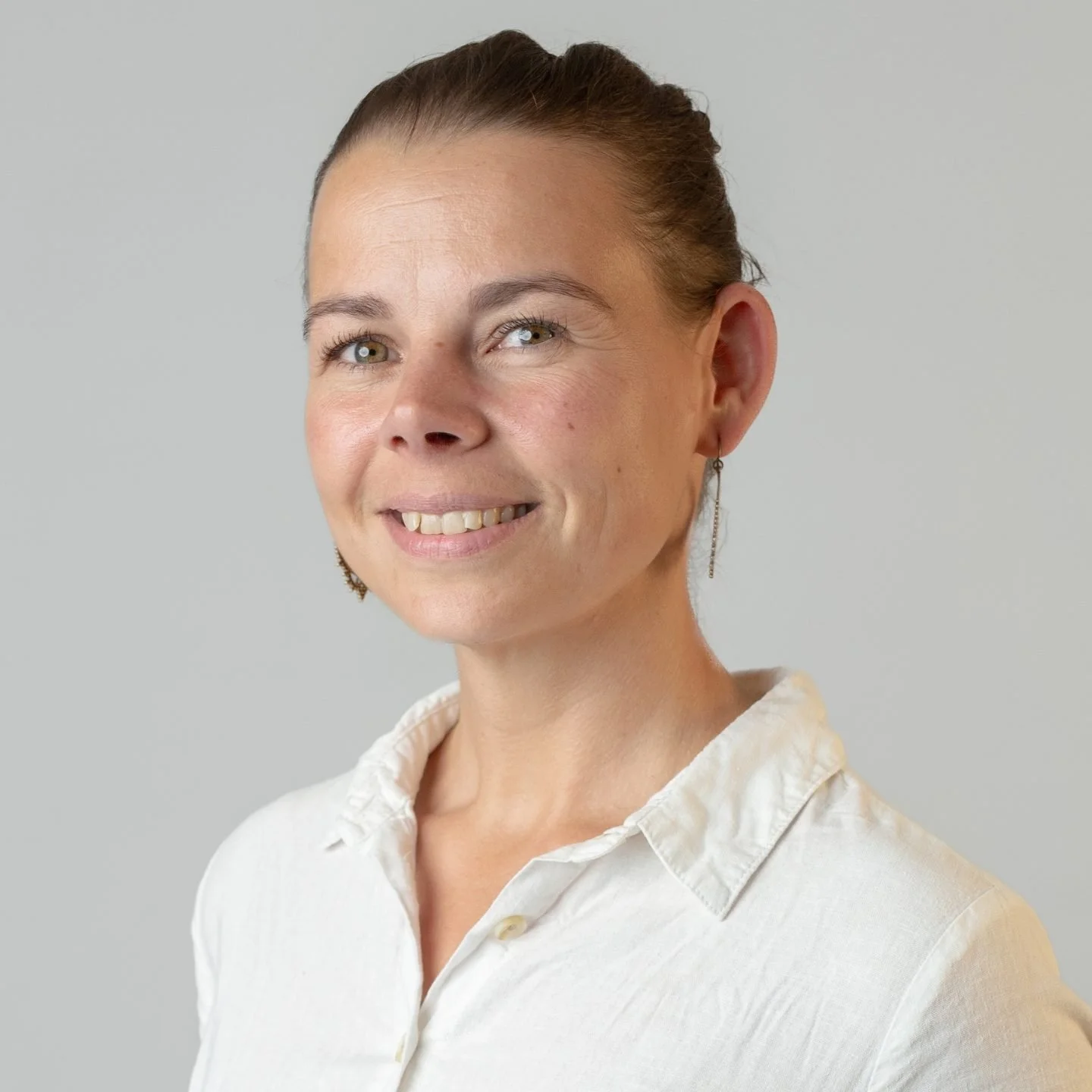 Portrait of a smiling woman with short brown hair and light-colored eyes, wearing a white collared shirt and dangling earrings, against a gray background.