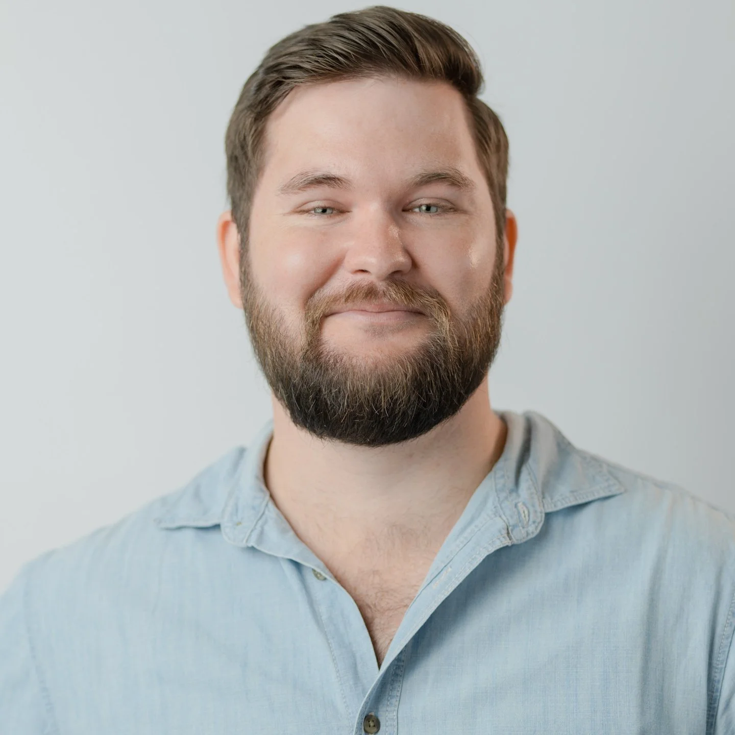 A smiling man with a full beard, light-colored hair, and blue eyes, wearing a light blue button-up shirt, against a plain white background.