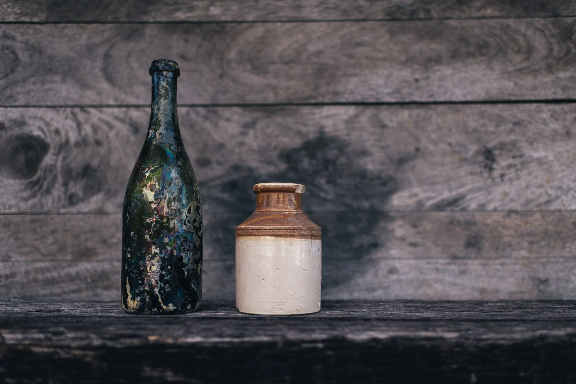 Three bottles, one with colorful paint splatters, another with a rustic brown and white finish, all arranged on a dark wooden surface against a wooden background.