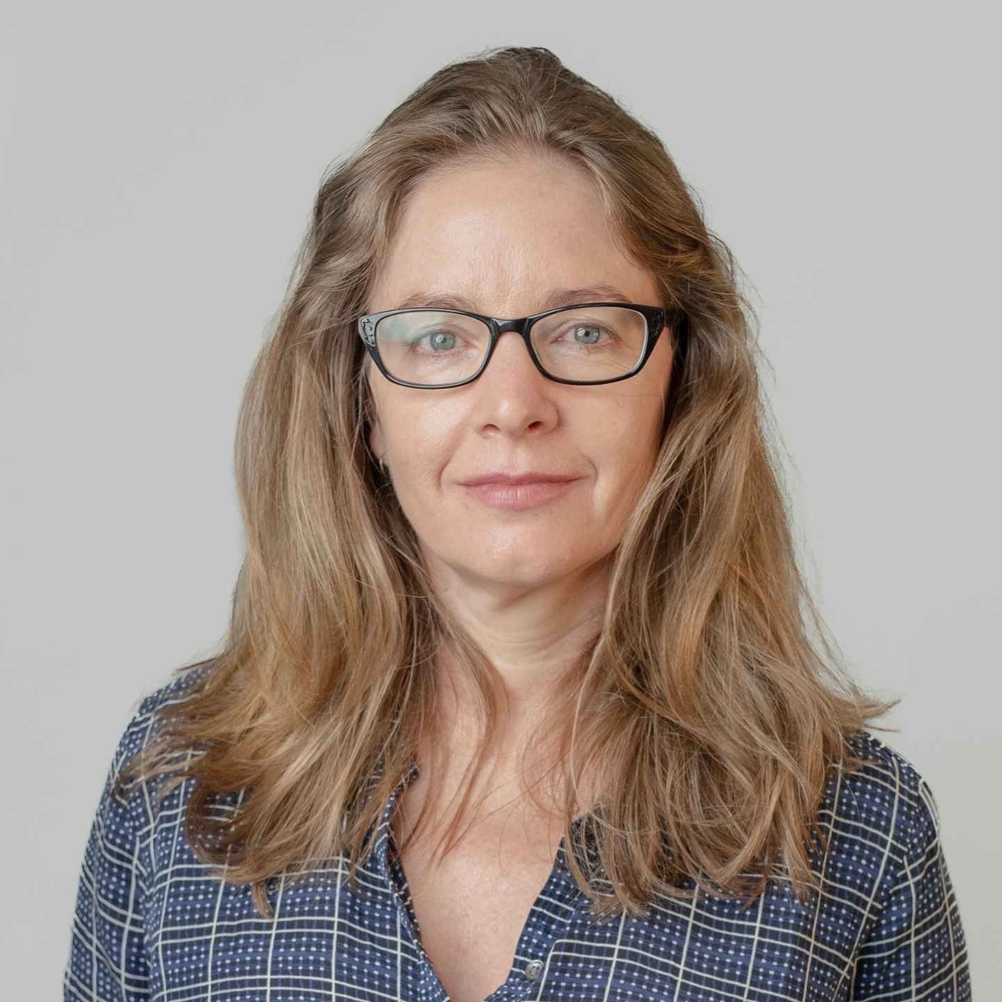 A woman with long wavy light brown hair wearing glasses and a navy blue checkered shirt, smiling softly against a plain gray background.