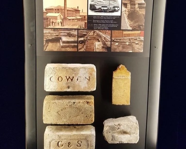 Historical display case with old bricks, one labeled 'COWEN' and another labeled 'C&S,' and an informational panel above showing photographs and text about a historical site.