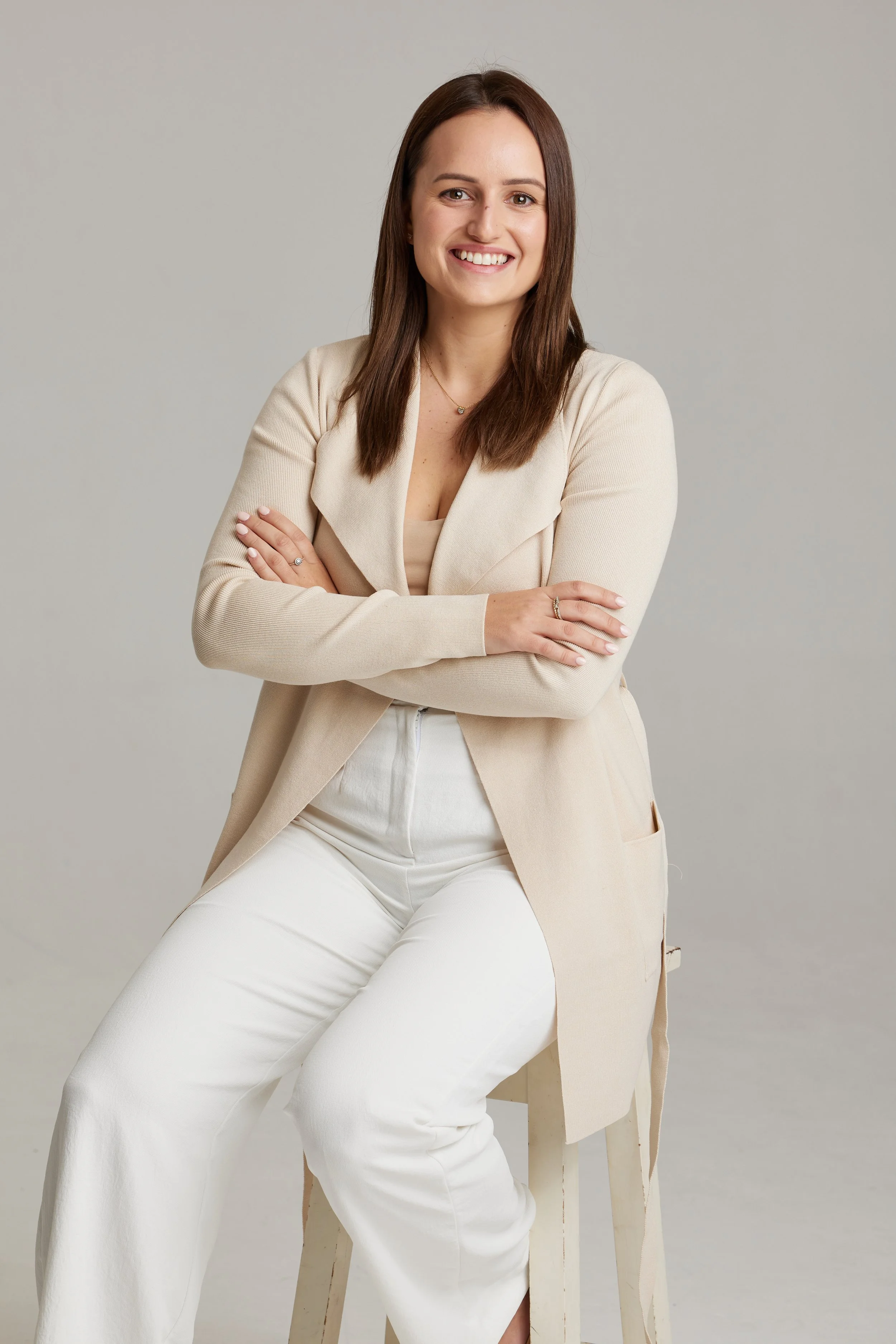 A woman sitting on a stool, smiling with crossed arms, wearing a beige blazer and white pants, against a plain neutral background.