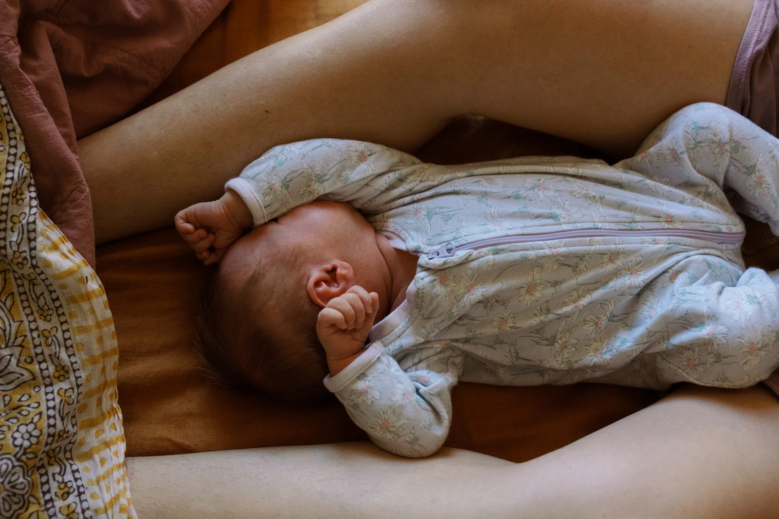 A newborn baby lying on a bed, dressed in a floral onesie, sleeping with hands near the face and a peaceful expression.