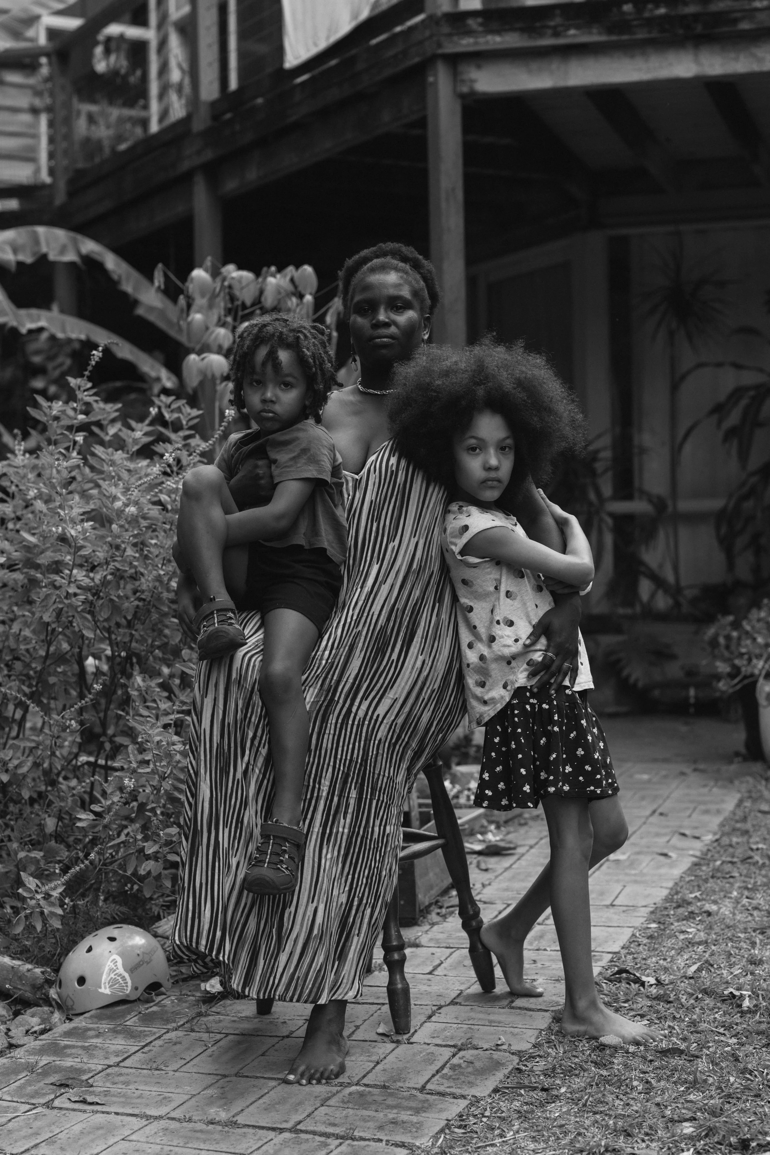 A woman and two children stand together in a backyard garden, with plants and a brick patio. The woman stands behind, holding a child on her lap, while a girl rests her arm on a chair and leans against the woman. The scene is in black and white.