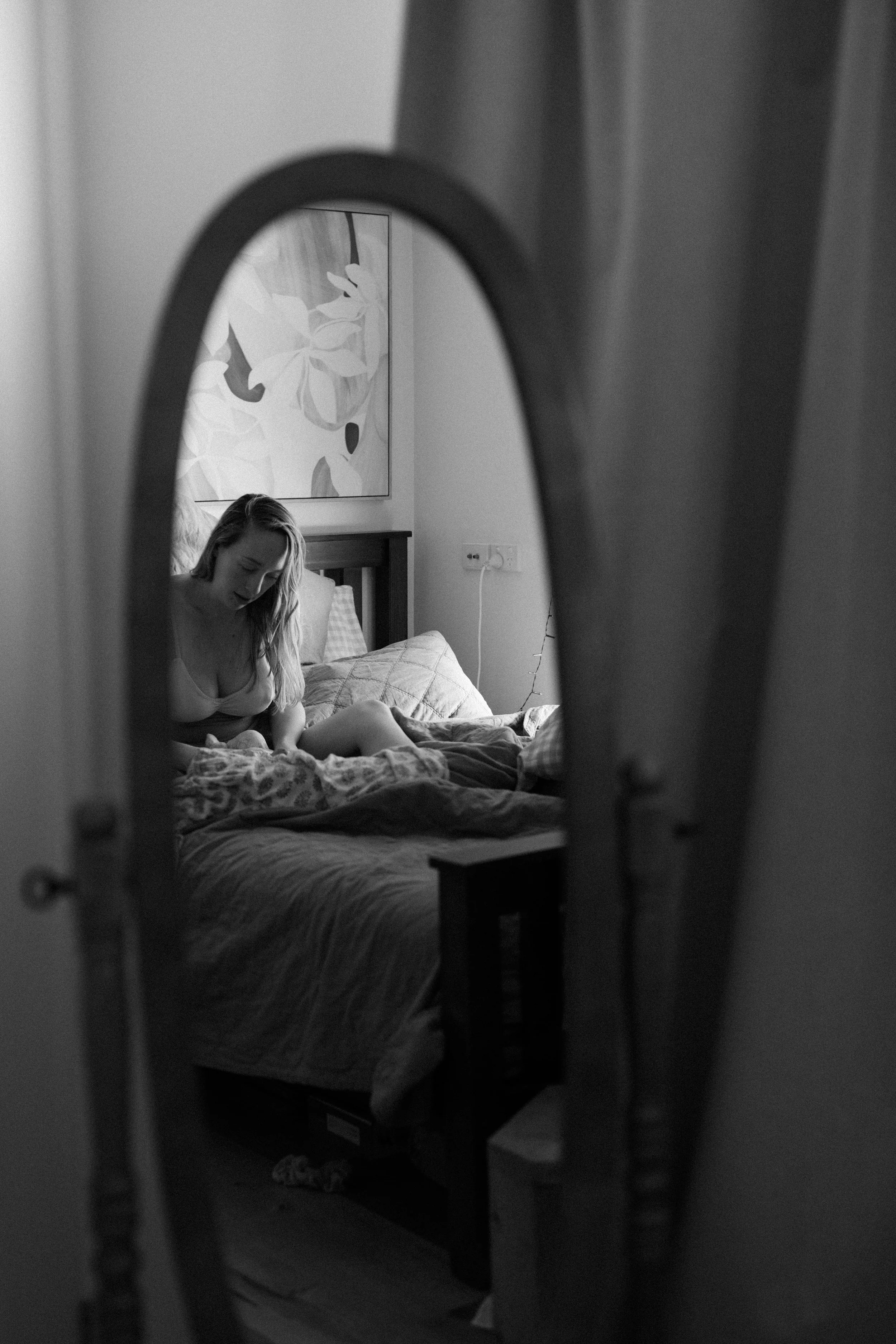A black and white photo of a woman sitting on a bed, viewed through a mirror or reflective surface, with a large abstract floral painting hanging on the wall behind her.