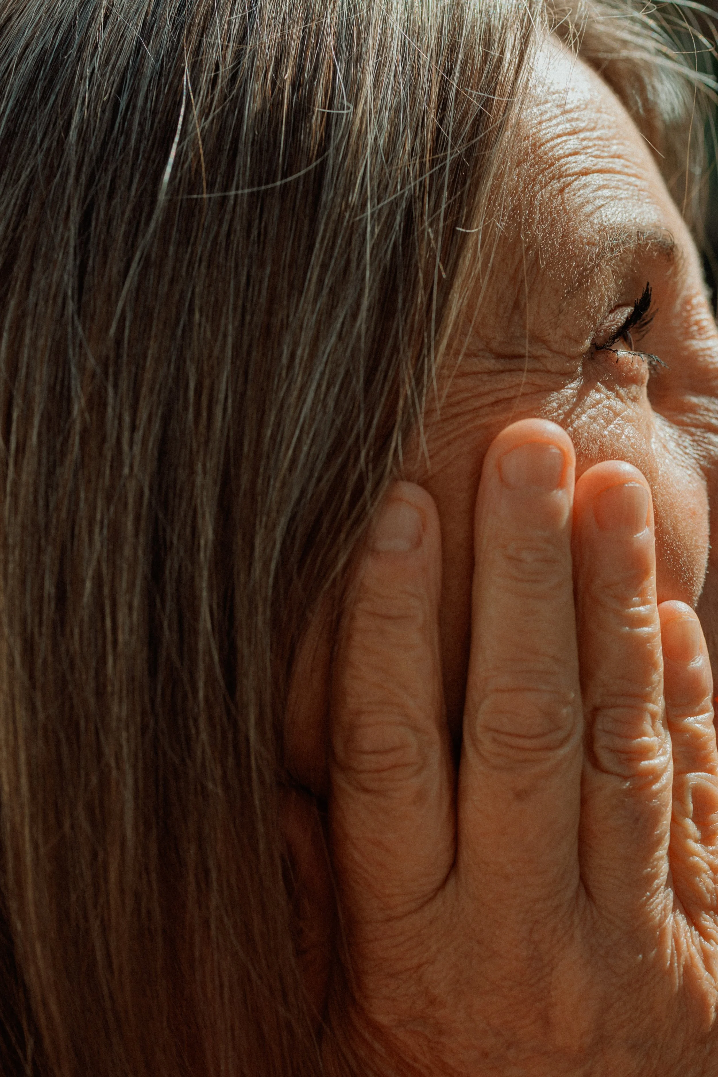 Close-up of a woman with long brown hair touching her face, focusing on her eye area and hand.