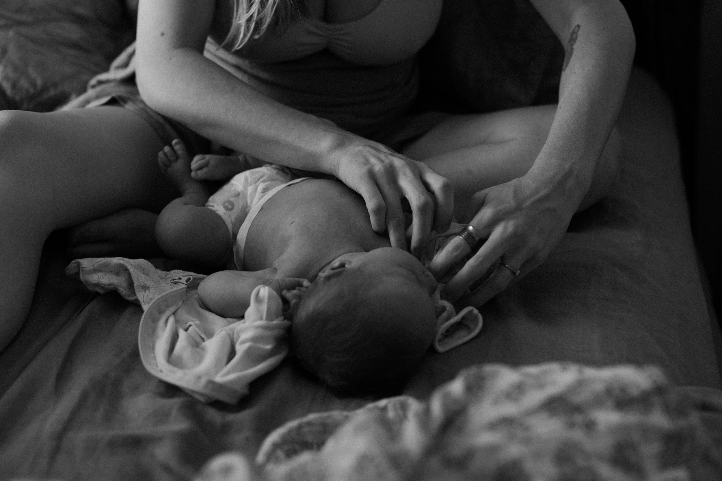 A woman breastfeeding an infant on a bed, with the baby lying on its back. The woman's hands are gently holding the baby's head and chest.