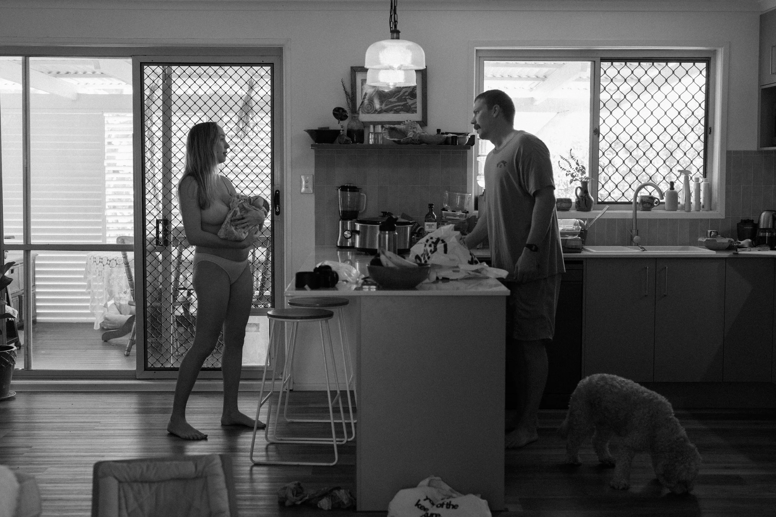 A woman holding a blanket in pajamas standing in the kitchen, talking to a man preparing food at the counter, with a dog on the floor nearby.