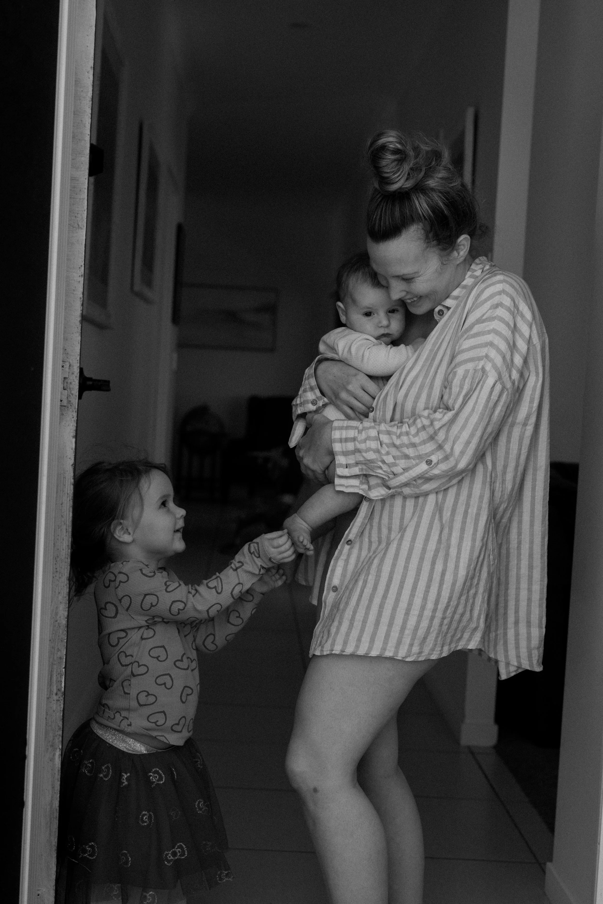 A woman holding a baby and a young girl reaching out to her at the doorway in a home.