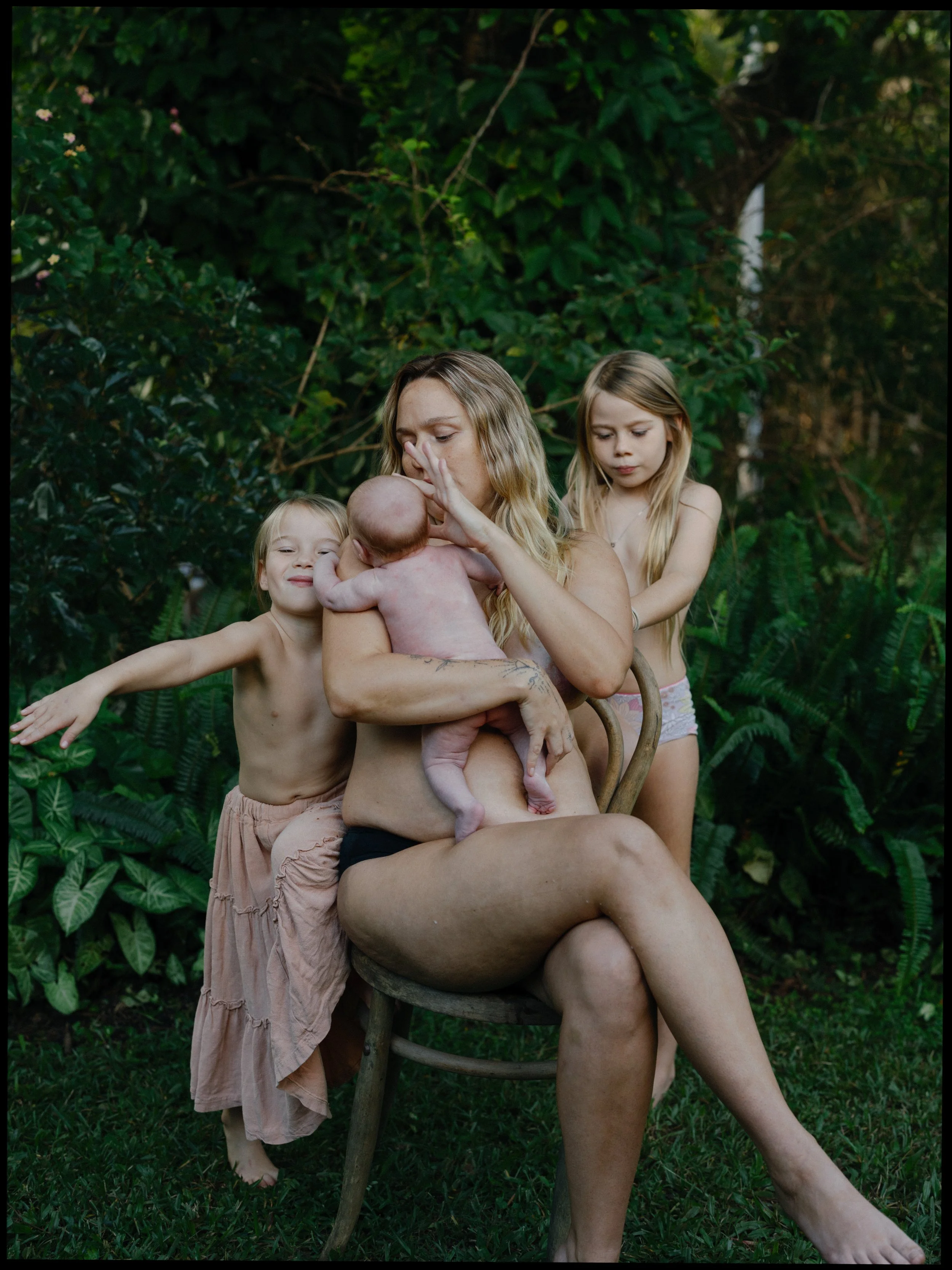 A woman holding a newborn baby with two young girls standing next to her outdoors surrounded by green foliage.
