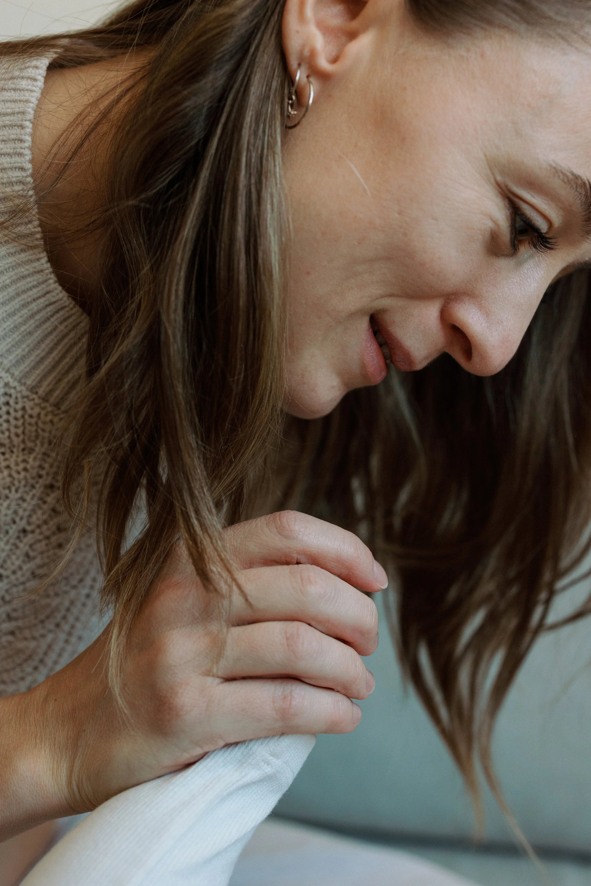 A woman with brown hair and earrings gently holding a white cloth or tissue, leaning forward with a soft smile.