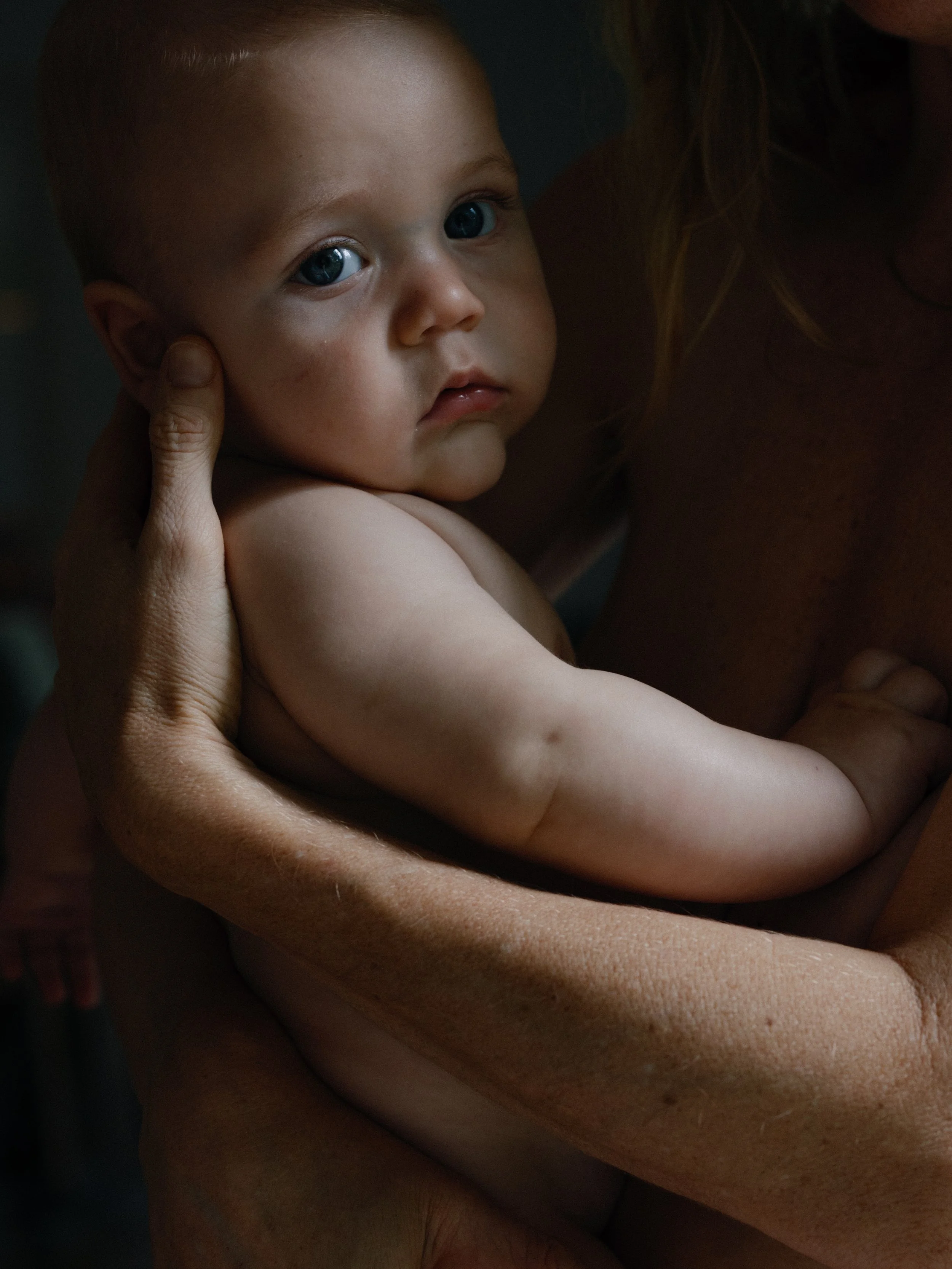 A young child with blue eyes being held gently by an adult, possibly a mother, with the child's face turned toward the camera and an expression of curiosity or concern.