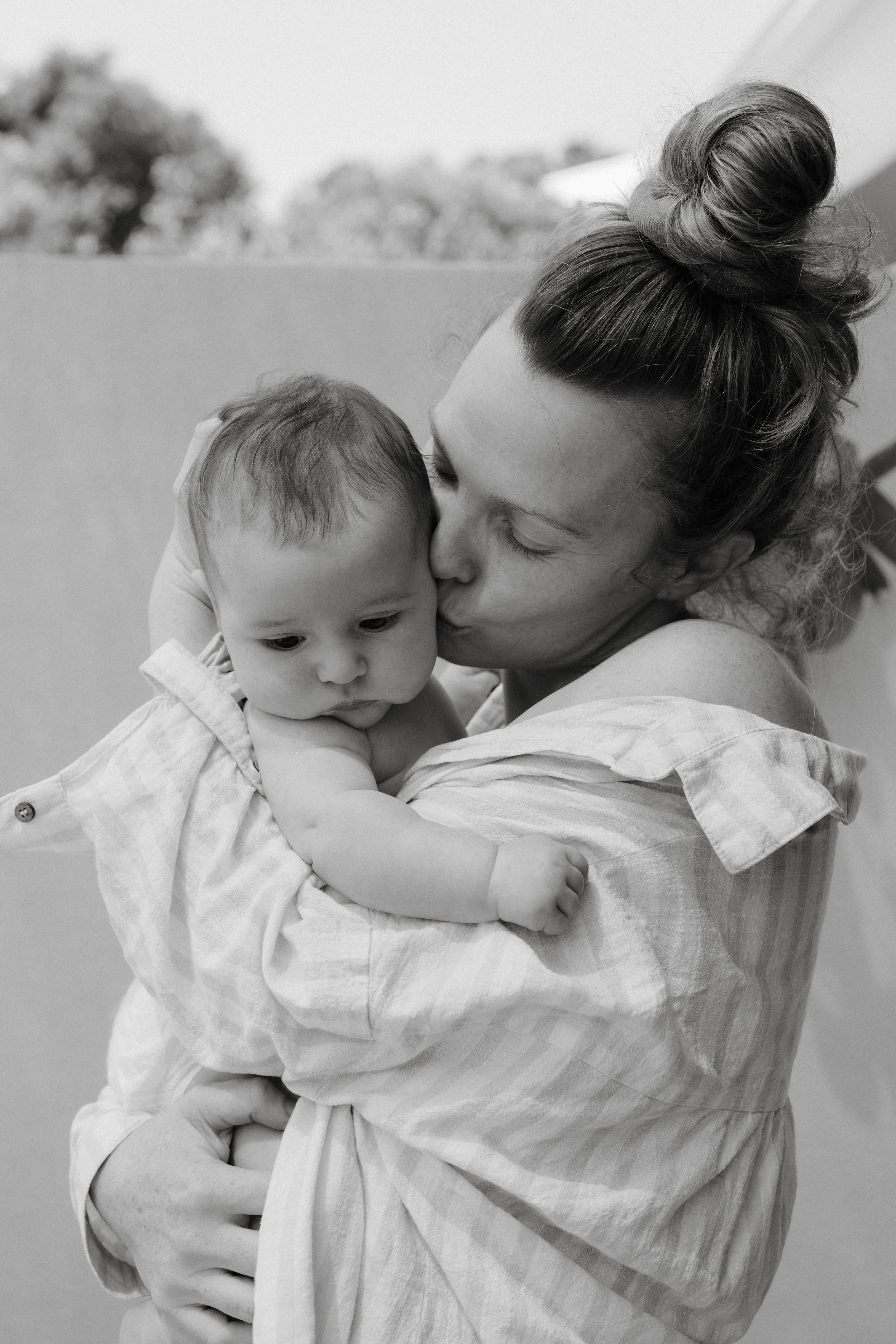A woman holding and kissing a baby on her shoulder outdoors.