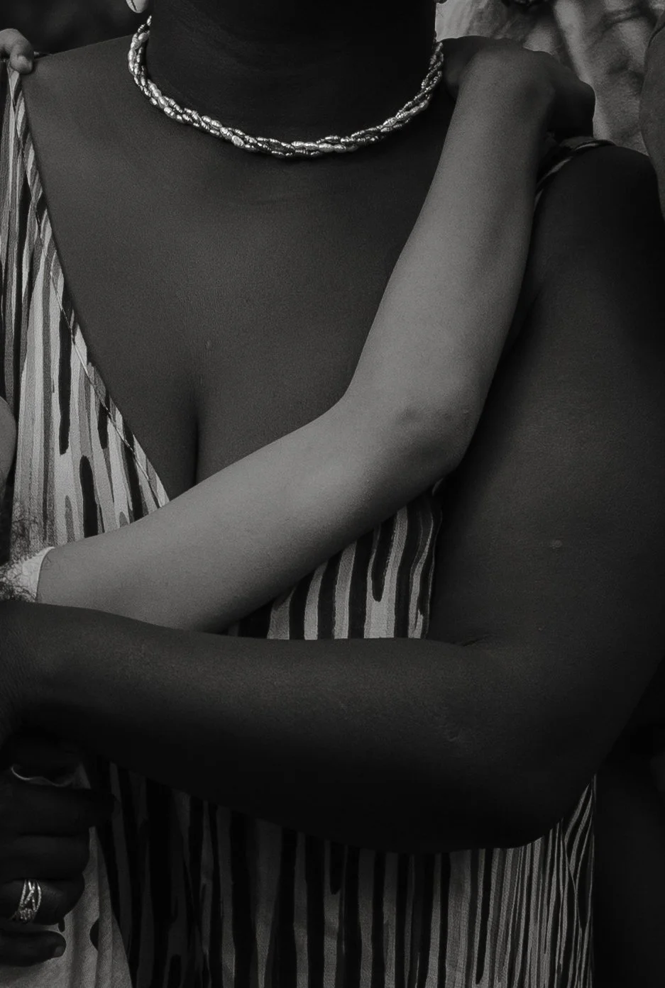 Close-up of two people embracing, showing parts of their arms, torsos, and necks. One person is wearing a striped garment and a beaded necklace.