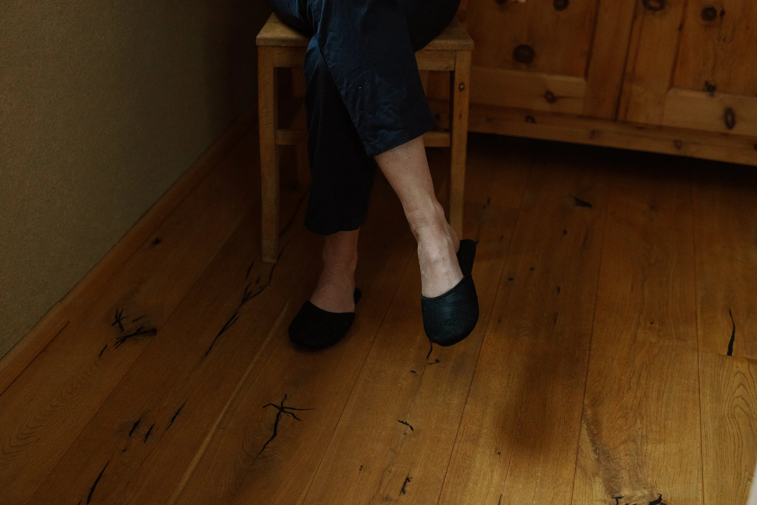 Close-up of a person's lower leg and foot, wearing a black shoe, lying on a wooden floor with black cracks or markings, next to a small wooden table and a wooden wall.