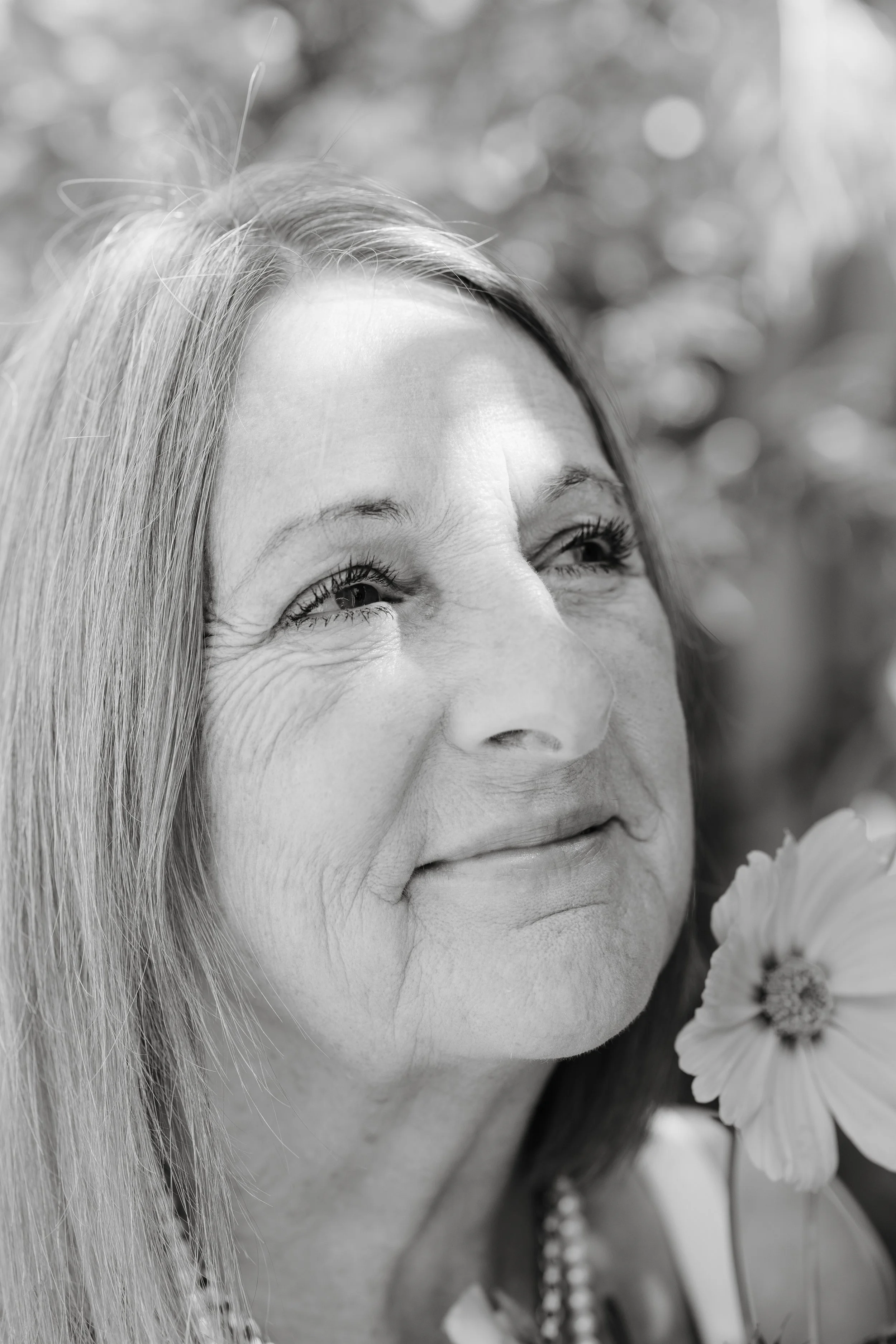 A close-up black and white photo of an elderly woman with long hair, smiling gently, with visible wrinkles, holding a flower near her face, in an outdoor setting with blurred background.