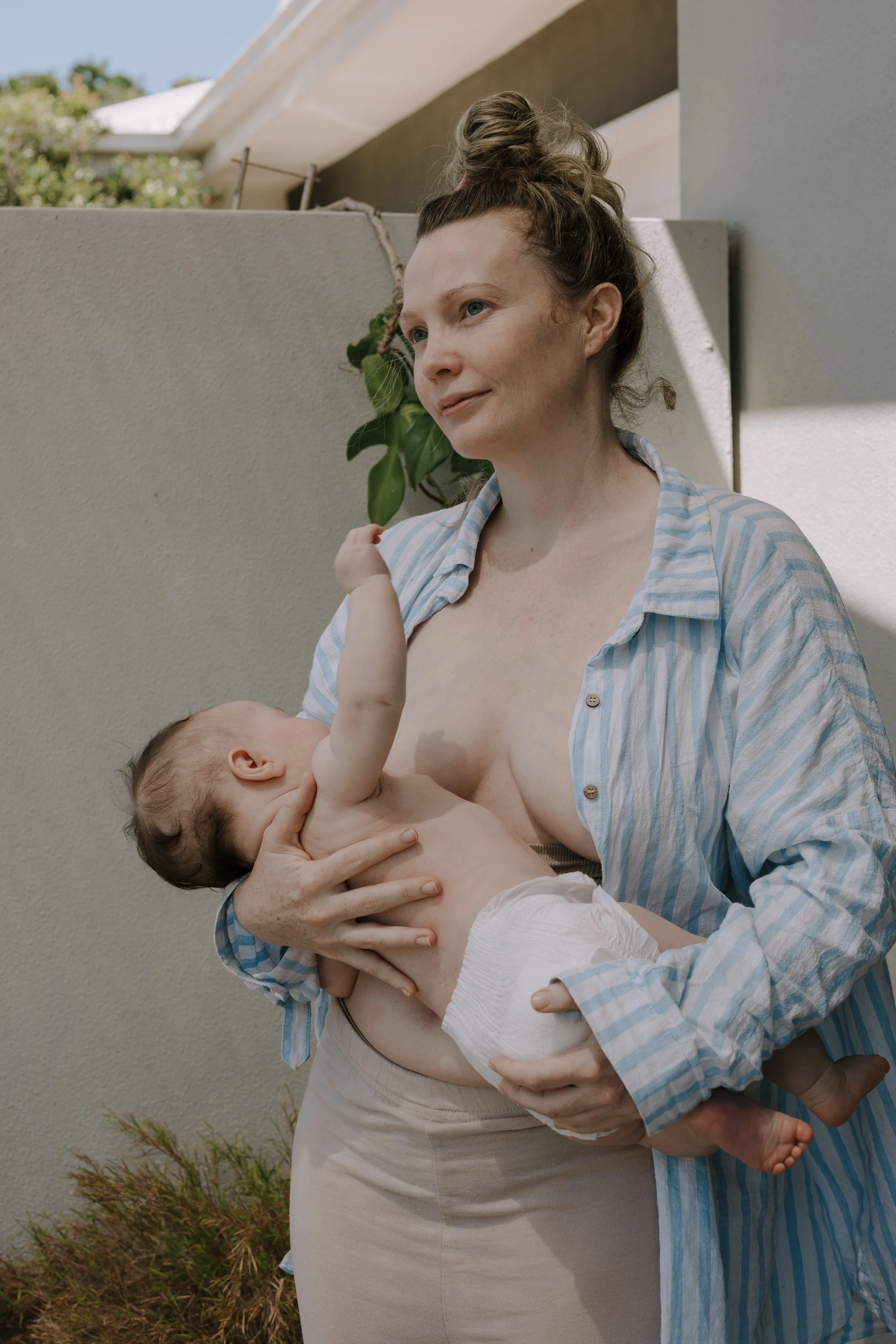 A woman with a topknot holding a baby outside in front of a beige wall, with a house and greenery in the background. The woman is wearing a blue and white striped shirt.