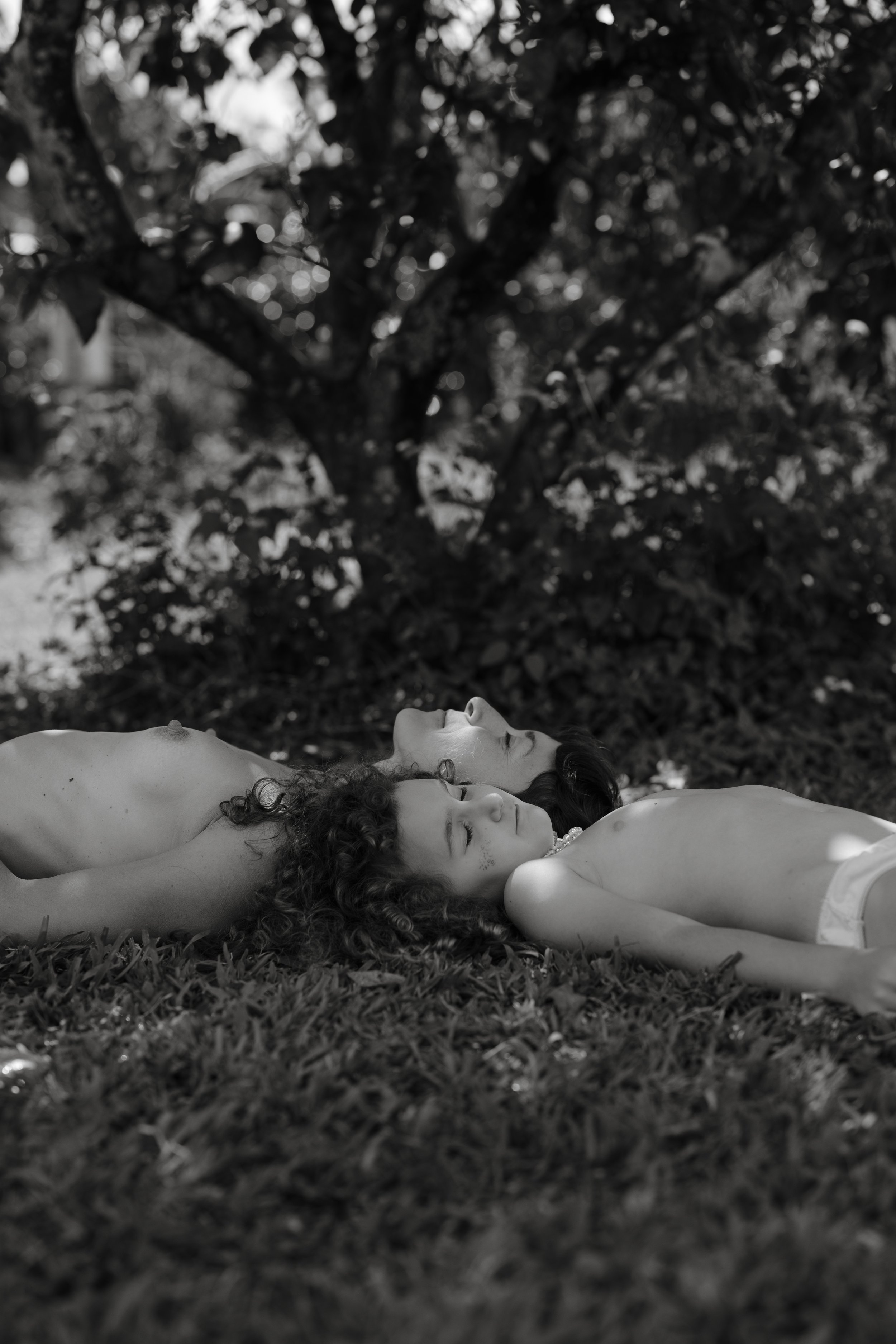 Two naked women lying on grass with eyes closed under a tree in a black-and-white photo.