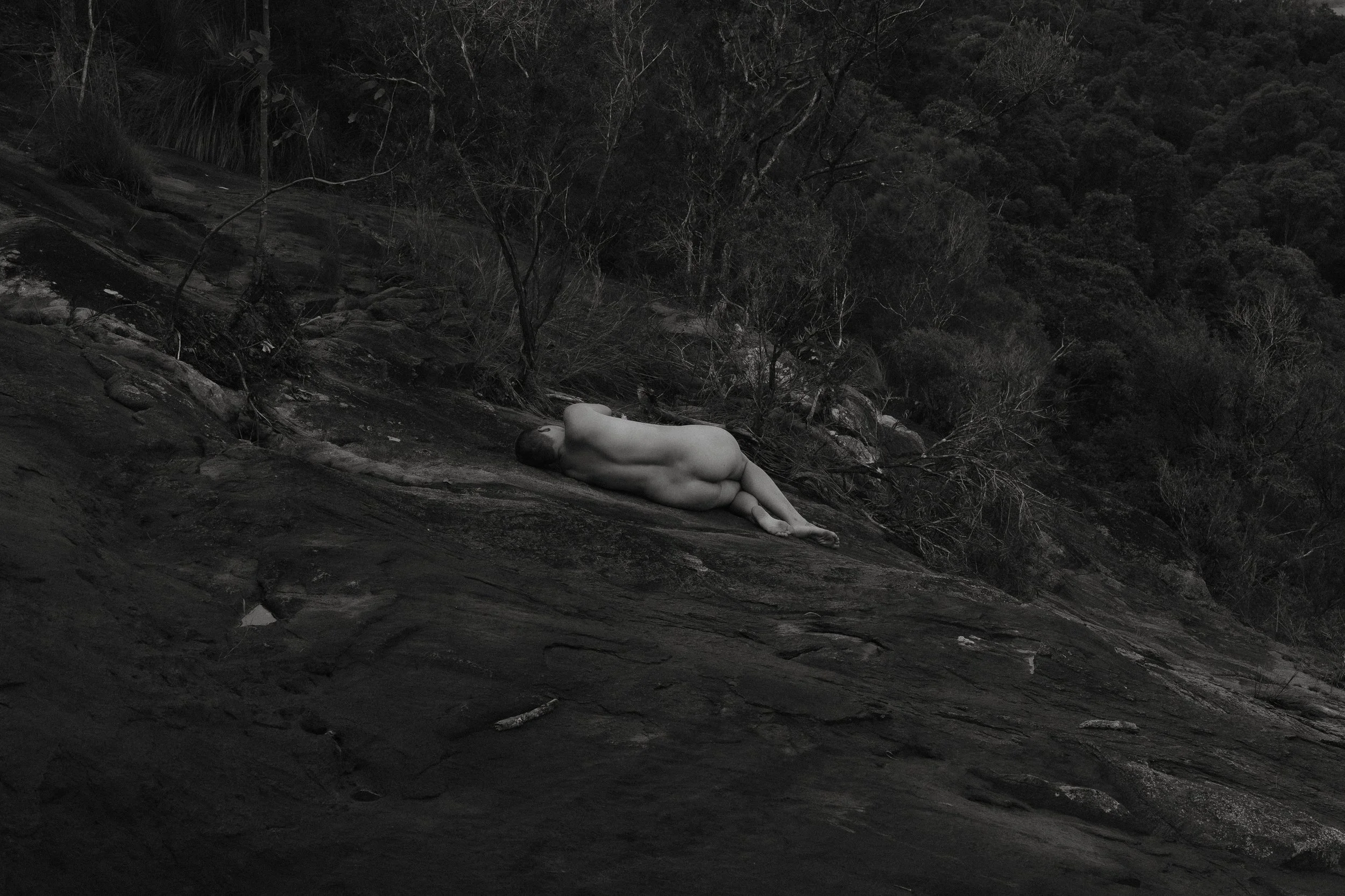 A woman lying on their side on a rocky hillside surrounded by sparse trees and bushes, in a natural outdoor setting.