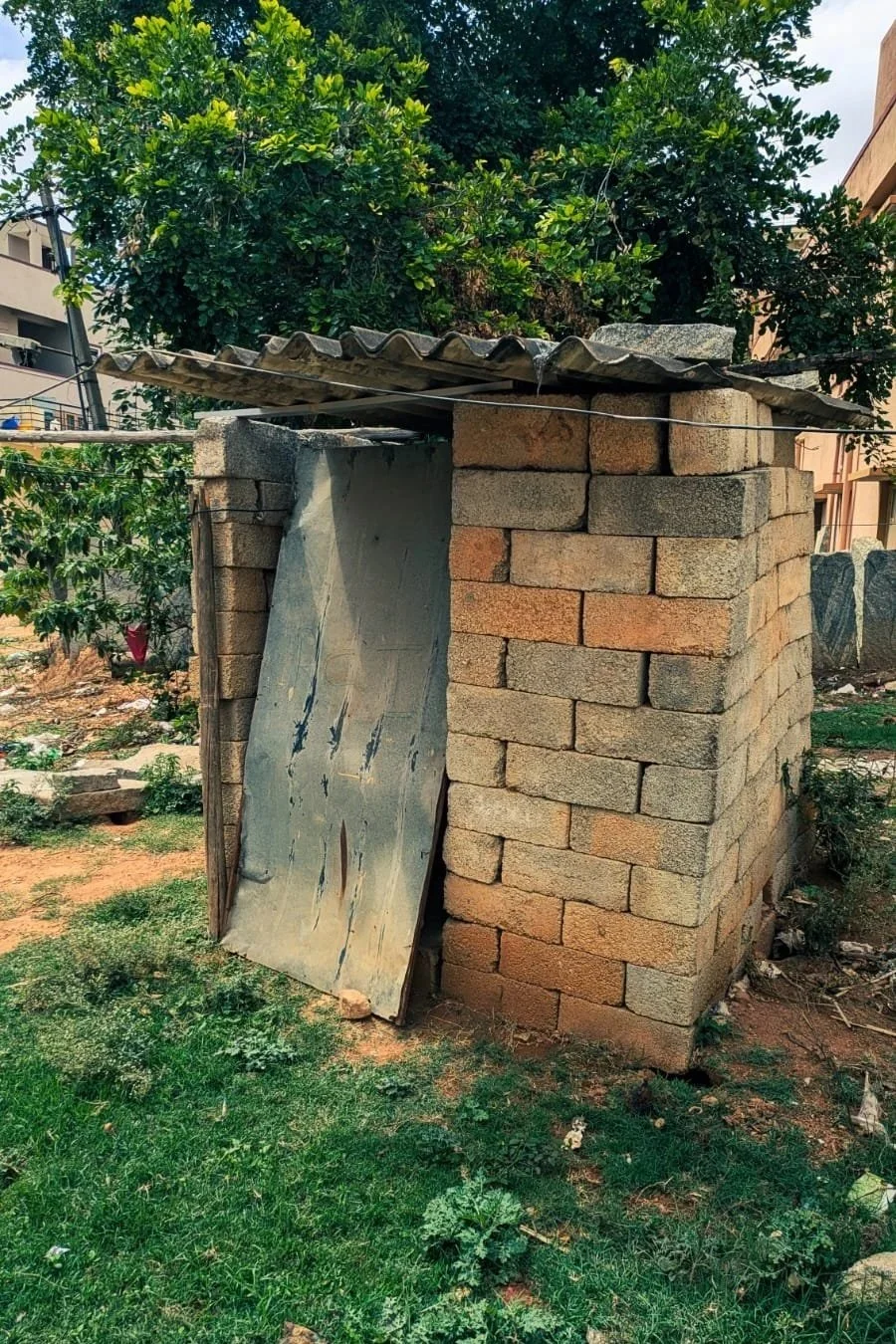 A small brick structure with a corrugated metal roof, leaning against the bricks, with a leafy tree in the background.