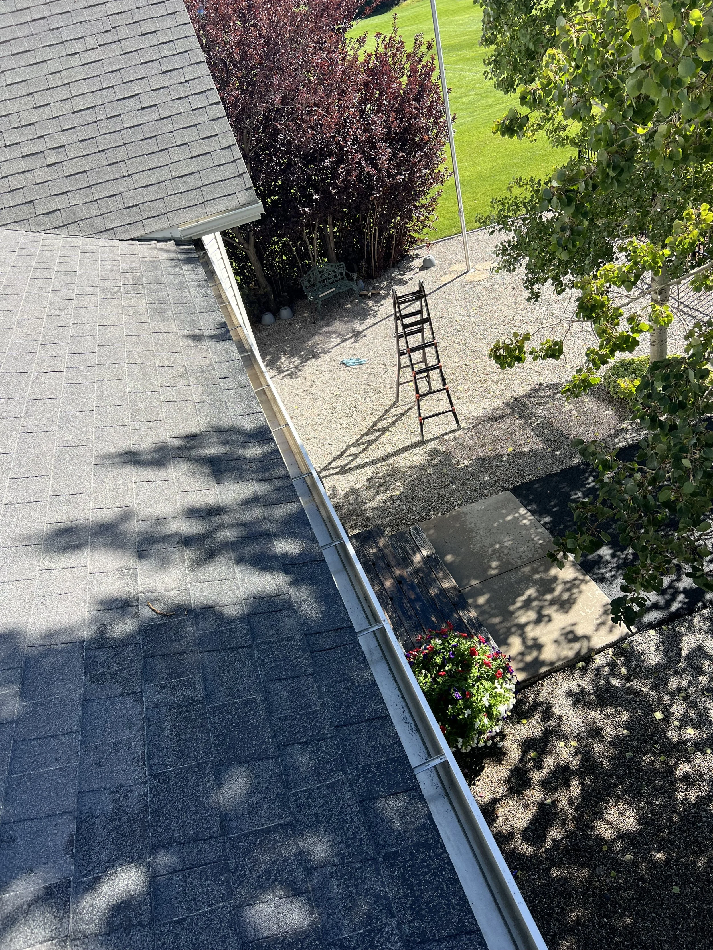 An aerial view of a backyard with a gravel area containing a ladder, a park bench, a flagpole, a flower pot, and trees, with a lawn and walkway in the background.