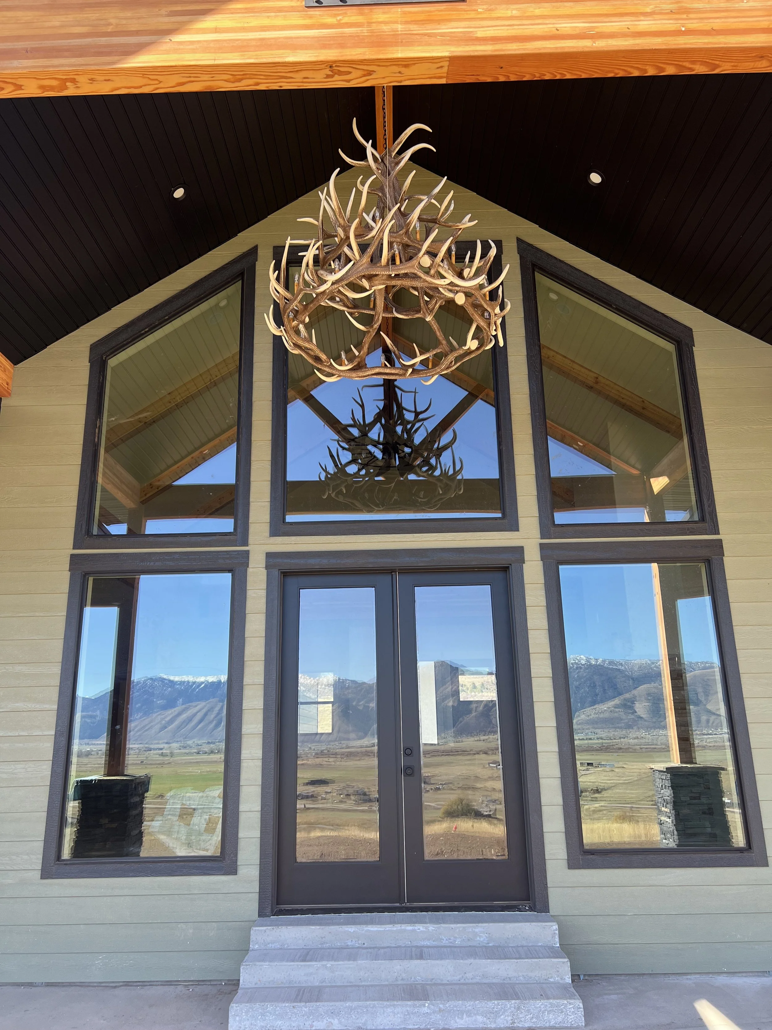 Front view of a modern house with large glass windows, a black door, and a rustic chandelier made of antlers hanging from the ceiling inside. The house has a mountain landscape visible through the windows.