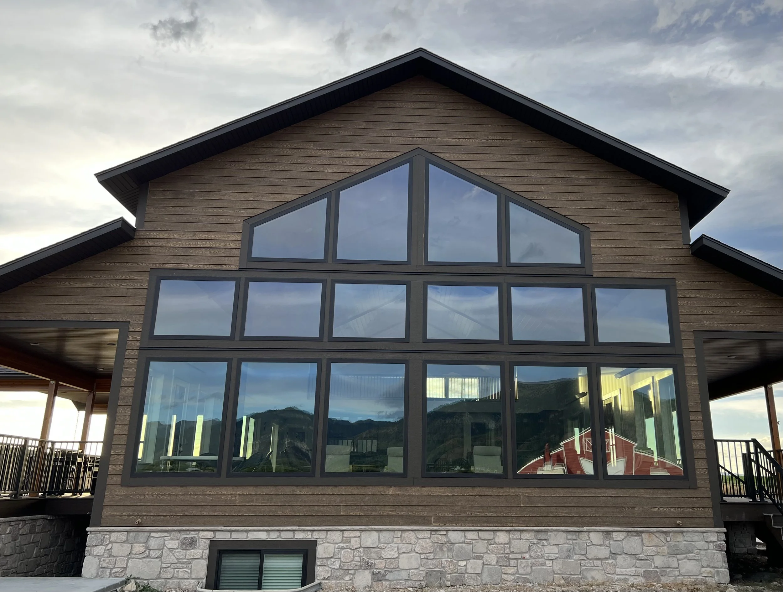 A multi-story house with a large set of floor-to-ceiling windows on the front, a stone foundation, gray trim, and a wood exterior. The house has a gabled roof and is set against a cloudy sky with mountains reflected in the windows.