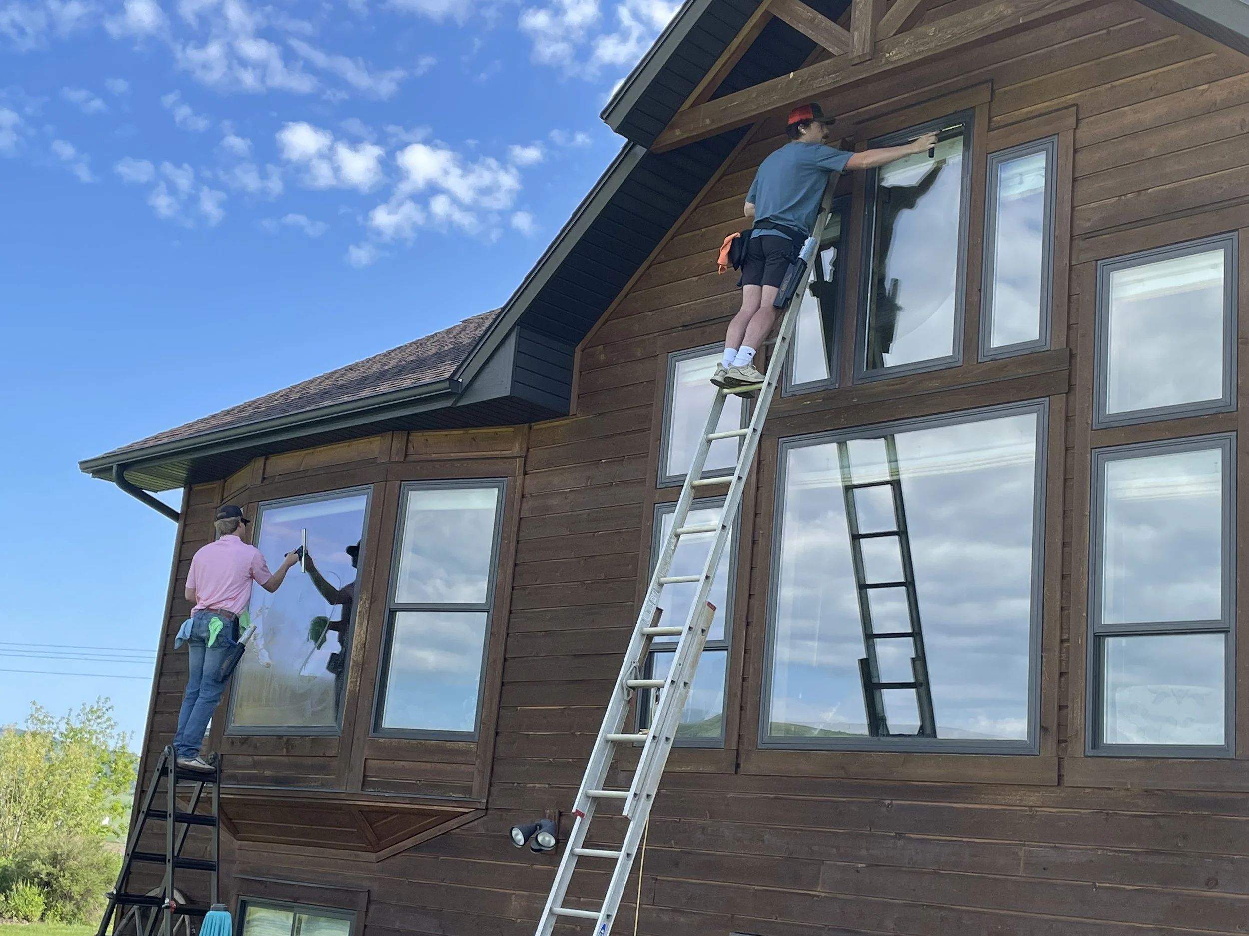 Two workers cleaning windows on a wooden house, one on a ladder and the other on the ground, with a blue sky and some clouds overhead.