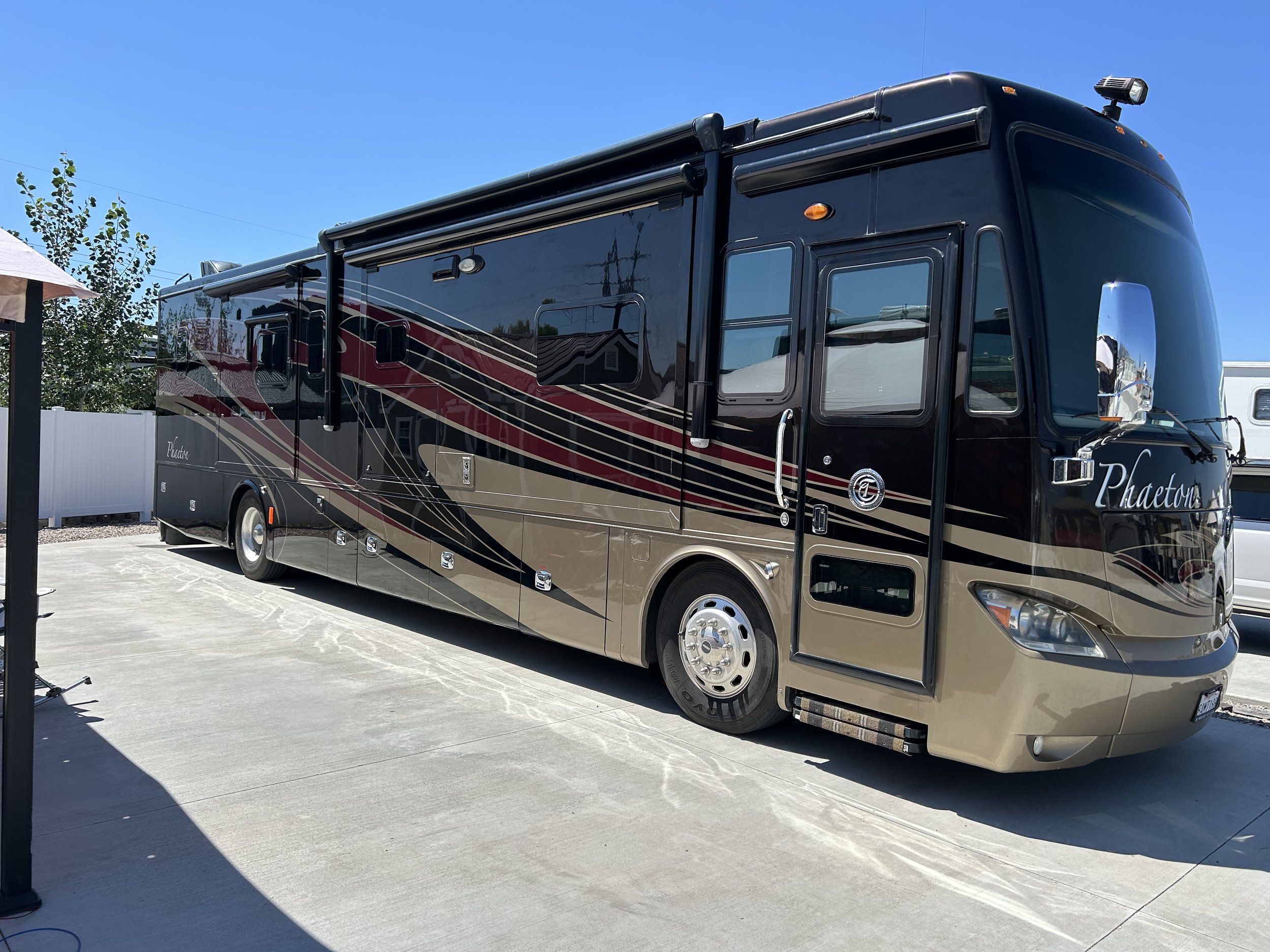 A black and tan Phaeton camper van with red and beige stripes parked on a concrete driveway under a clear blue sky.