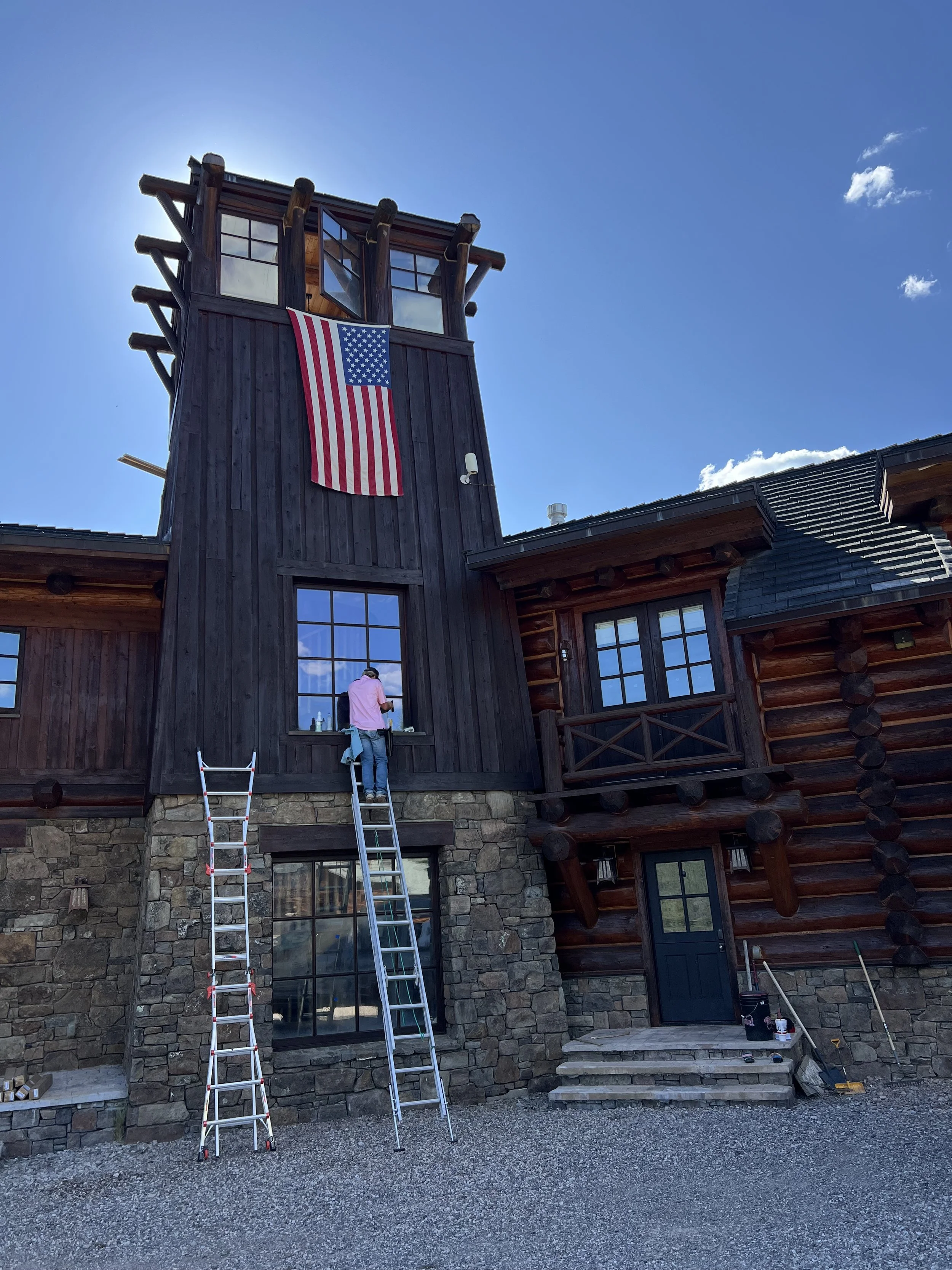 A person on a ladder cleaning a window on a large wooden building with a tall tower, an American flag hanging from the tower, and a blue sky in the background.