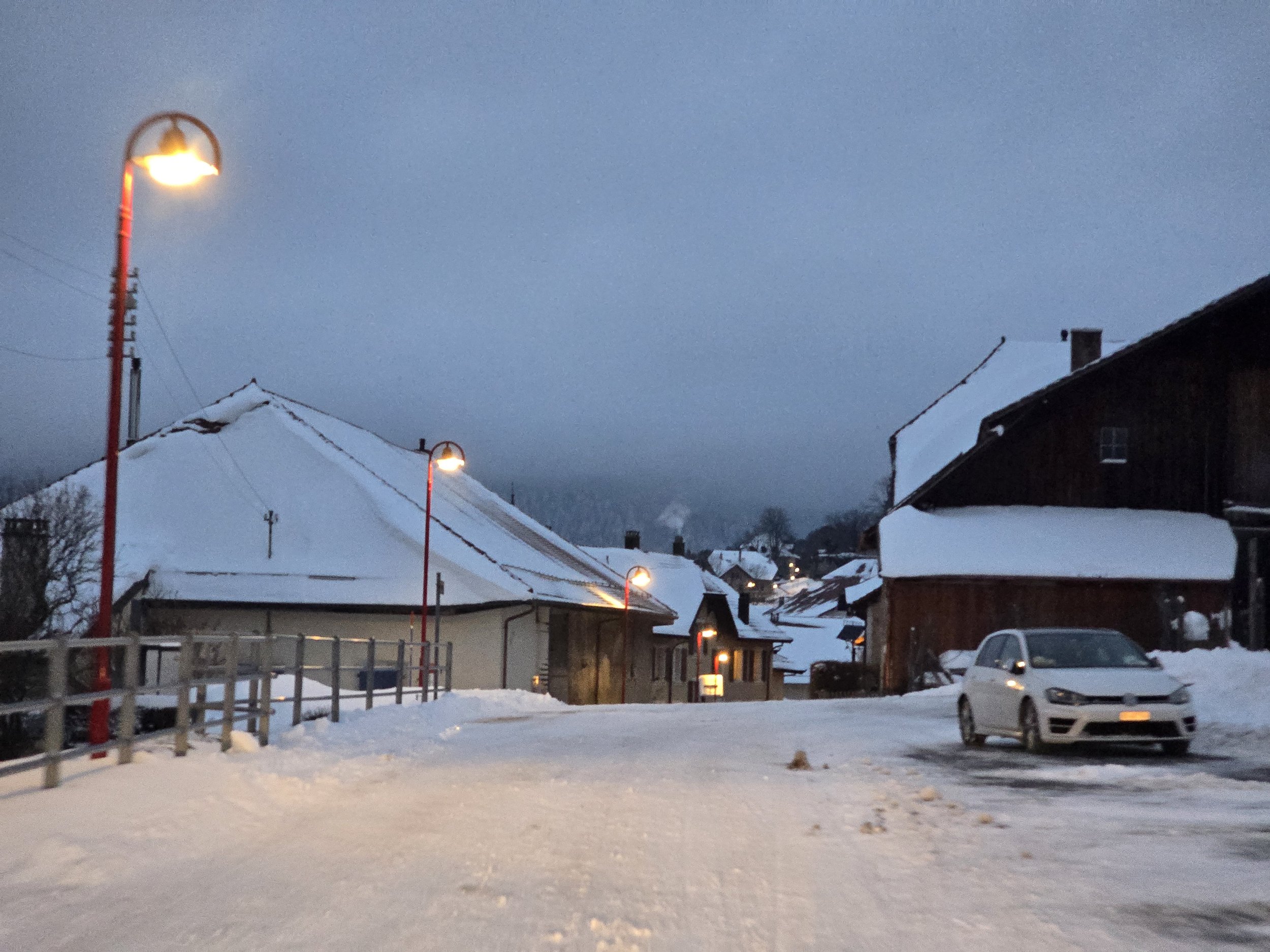 Las farolas despiertan el camino, como si cada luz fuera una historia que se niega a apagarse. Las casas, cubiertas de nieve, guardan el calor de quienes resisten al frío, y el cielo, pesado, observa en silencio.