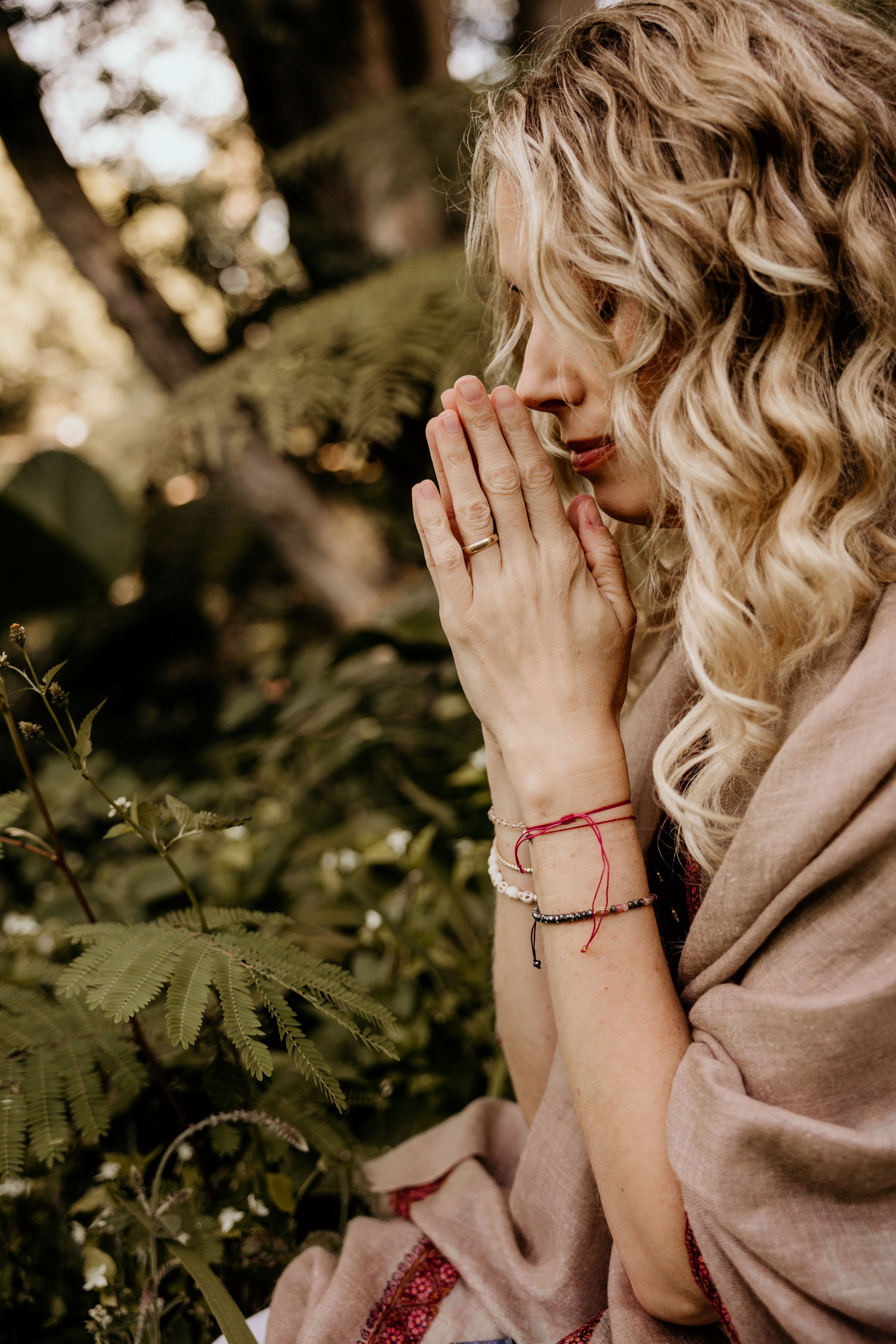 A woman with curly blonde hair, eyes closed, holding her hands in a prayer position near her face in a peaceful outdoor setting surrounded by greenery.