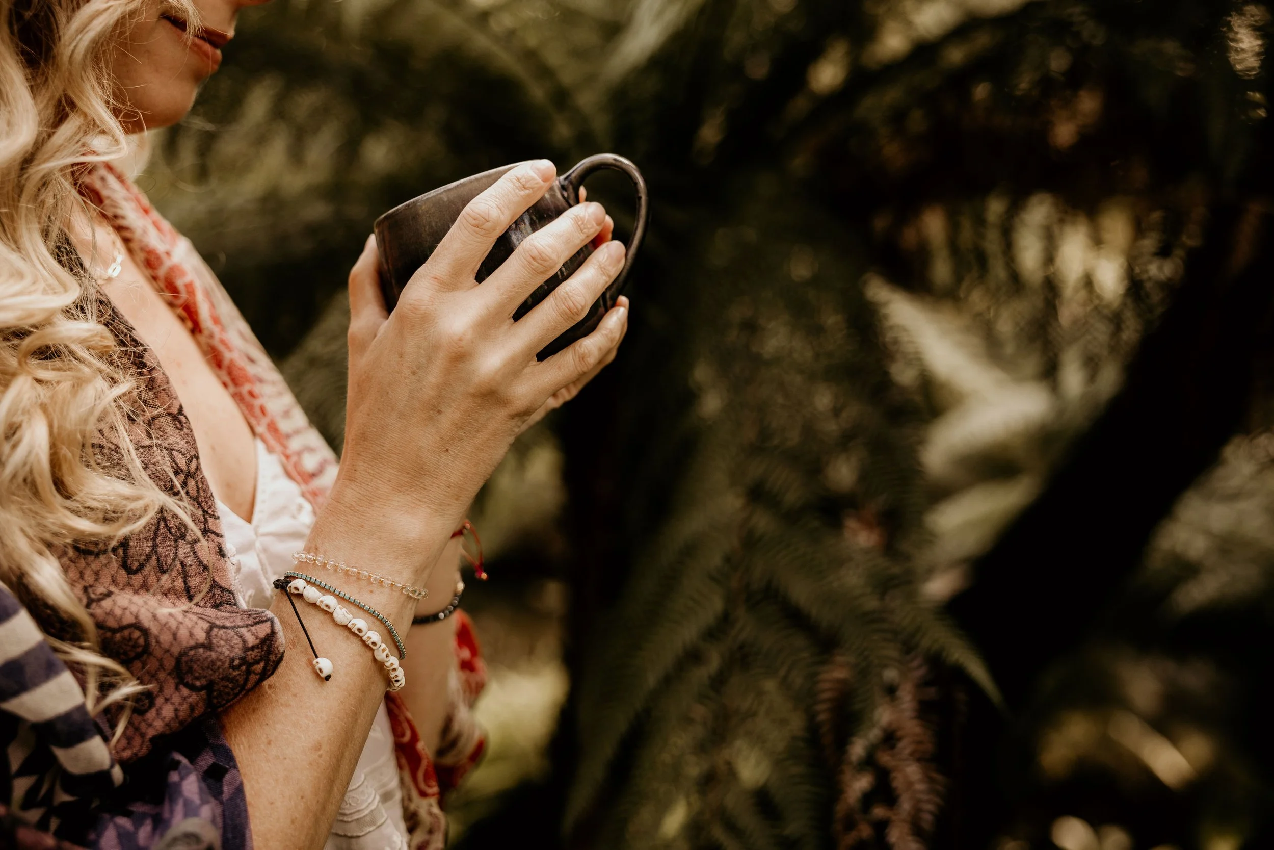 A woman with blonde curly hair holding a dark brown mug, standing outdoors with blurred trees and foliage in the background.