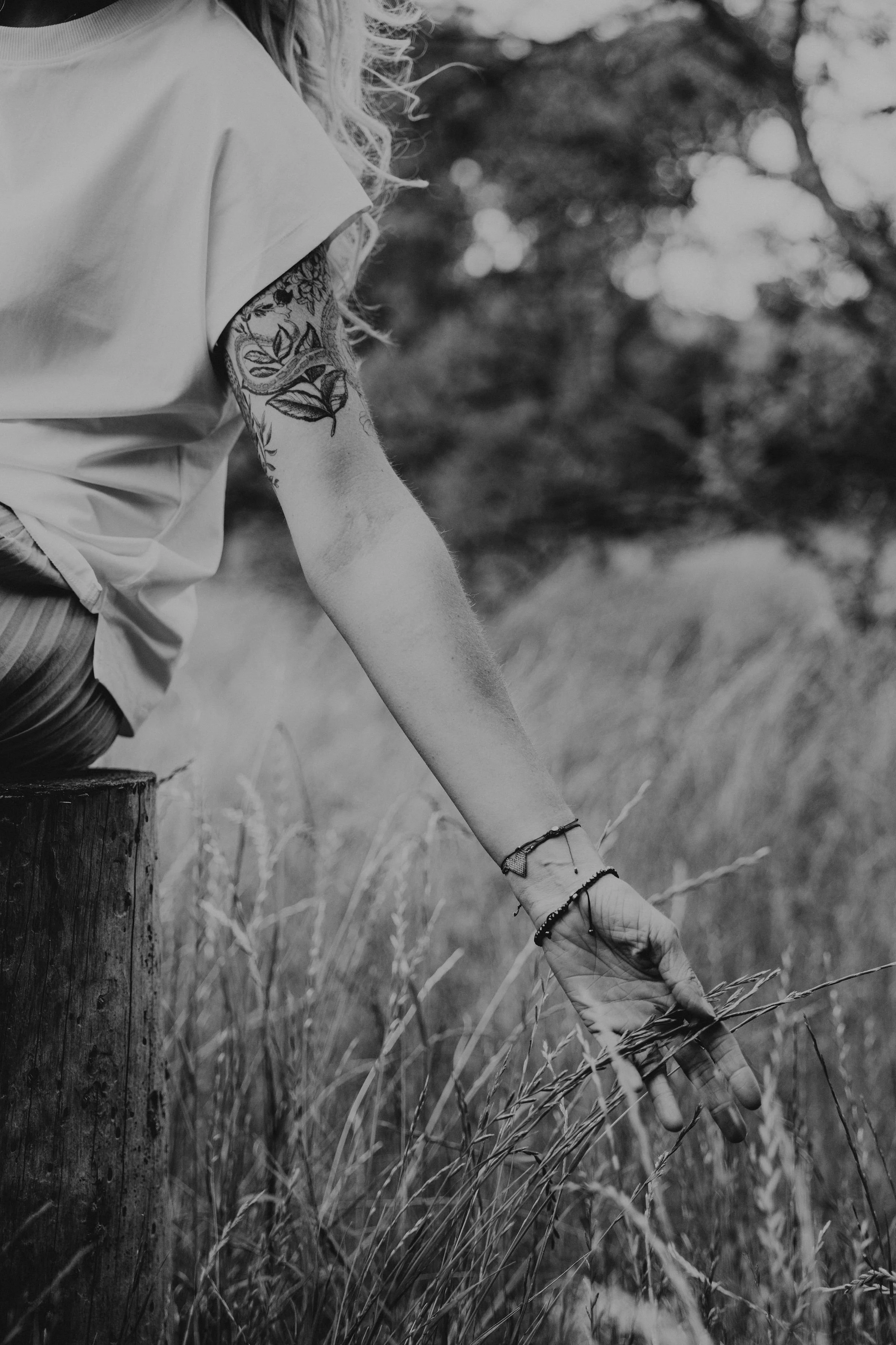 A person with a tattoo on their arm, wearing bracelets, sitting on a tree stump reaching down to touch tall grass in a natural outdoor setting, black and white photo.