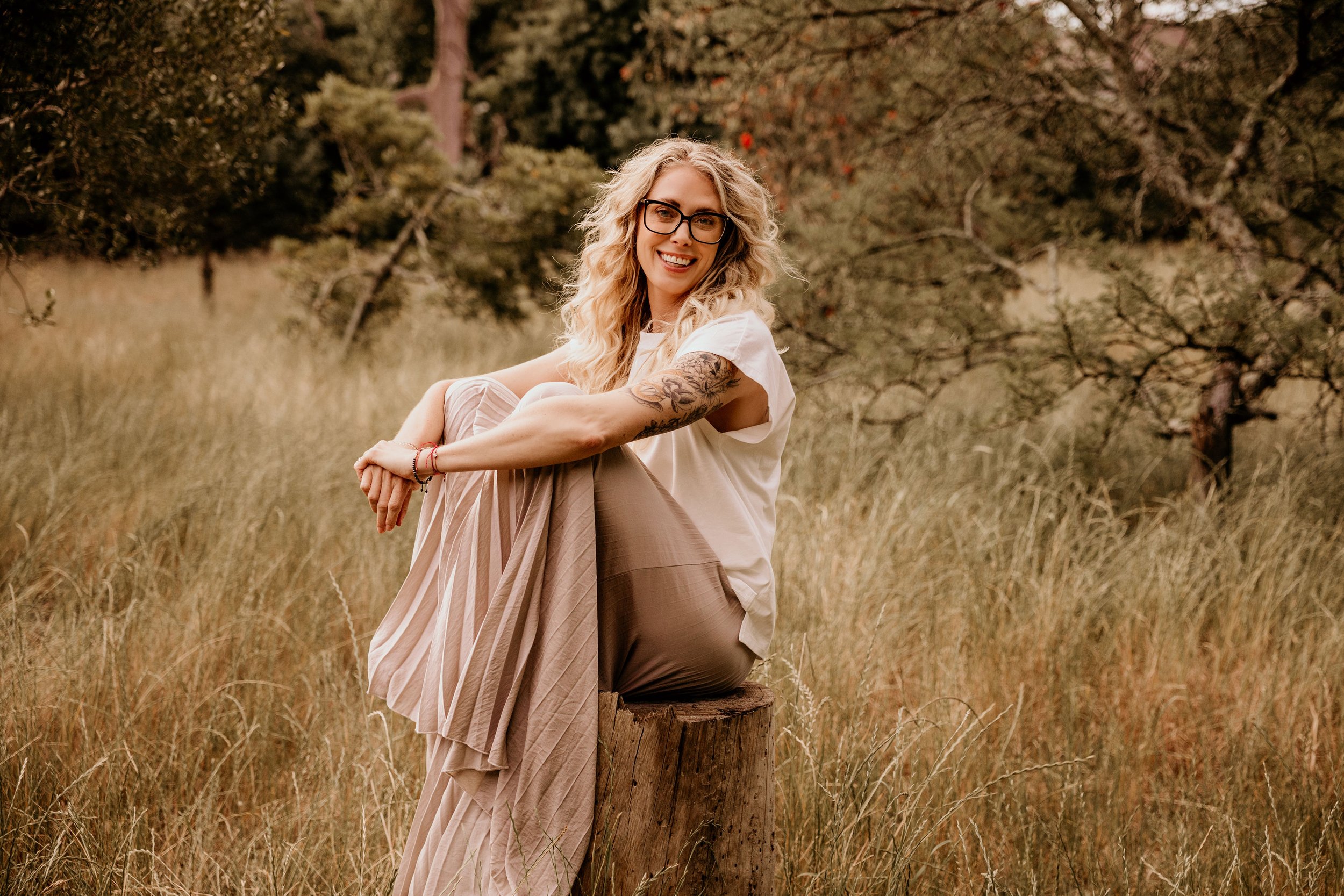 A young woman with curly blonde hair, glasses, and a tattoo on her arm, sitting on a tree stump in a grassy field, smiling at the camera.
