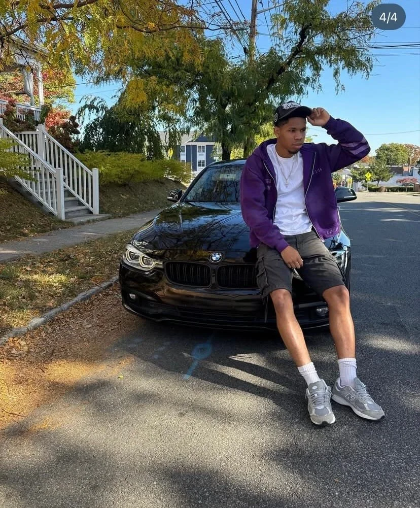 Young man in a purple jacket, white T-shirt, gray shorts, white socks, and sneakers sitting on the front of a black BMW parked on the street.