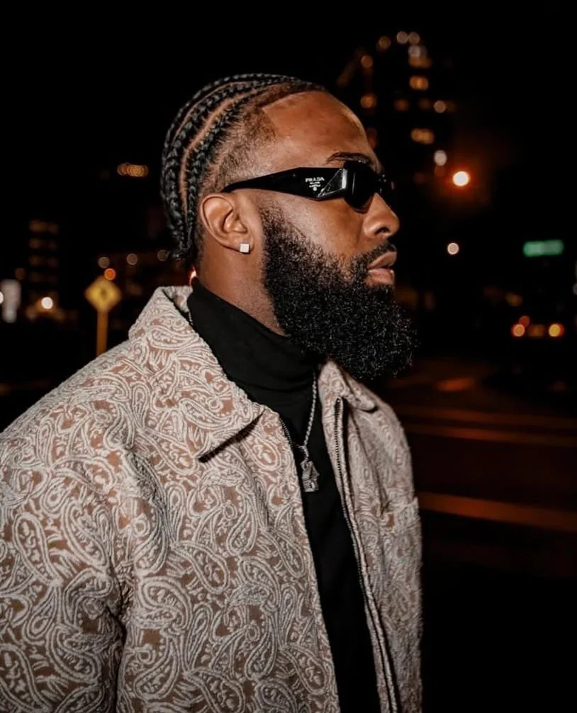 Profile of a man with braided hair, beard, black Prada sunglasses, diamond earrings, and a patterned beige jacket at night.