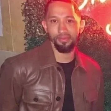 A man with a beard and mustache wearing a brown leather jacket standing outdoors at night.