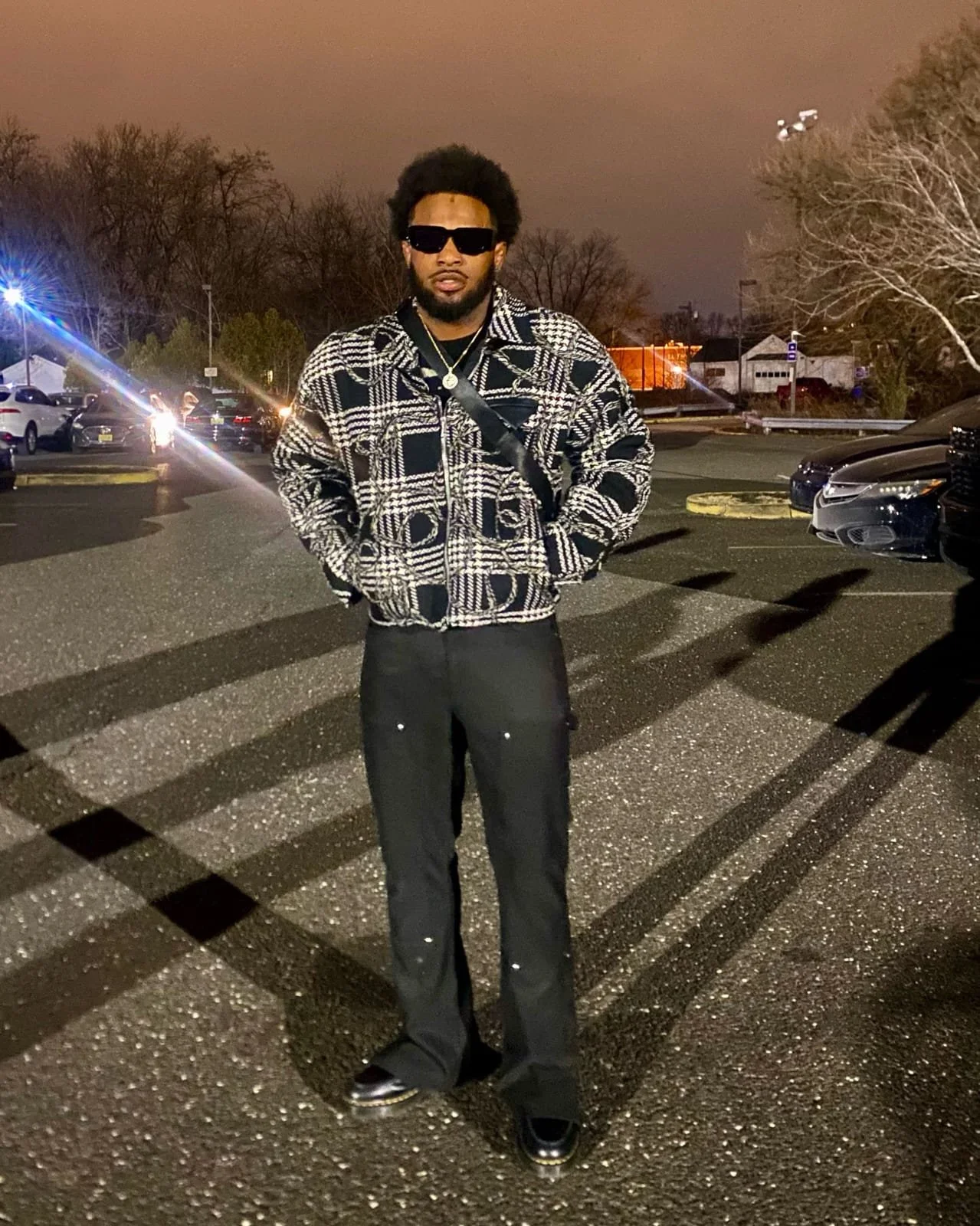 A man standing in a parking lot at night, wearing sunglasses, a patterned jacket, black pants, and black shoes, with cars and trees in the background.