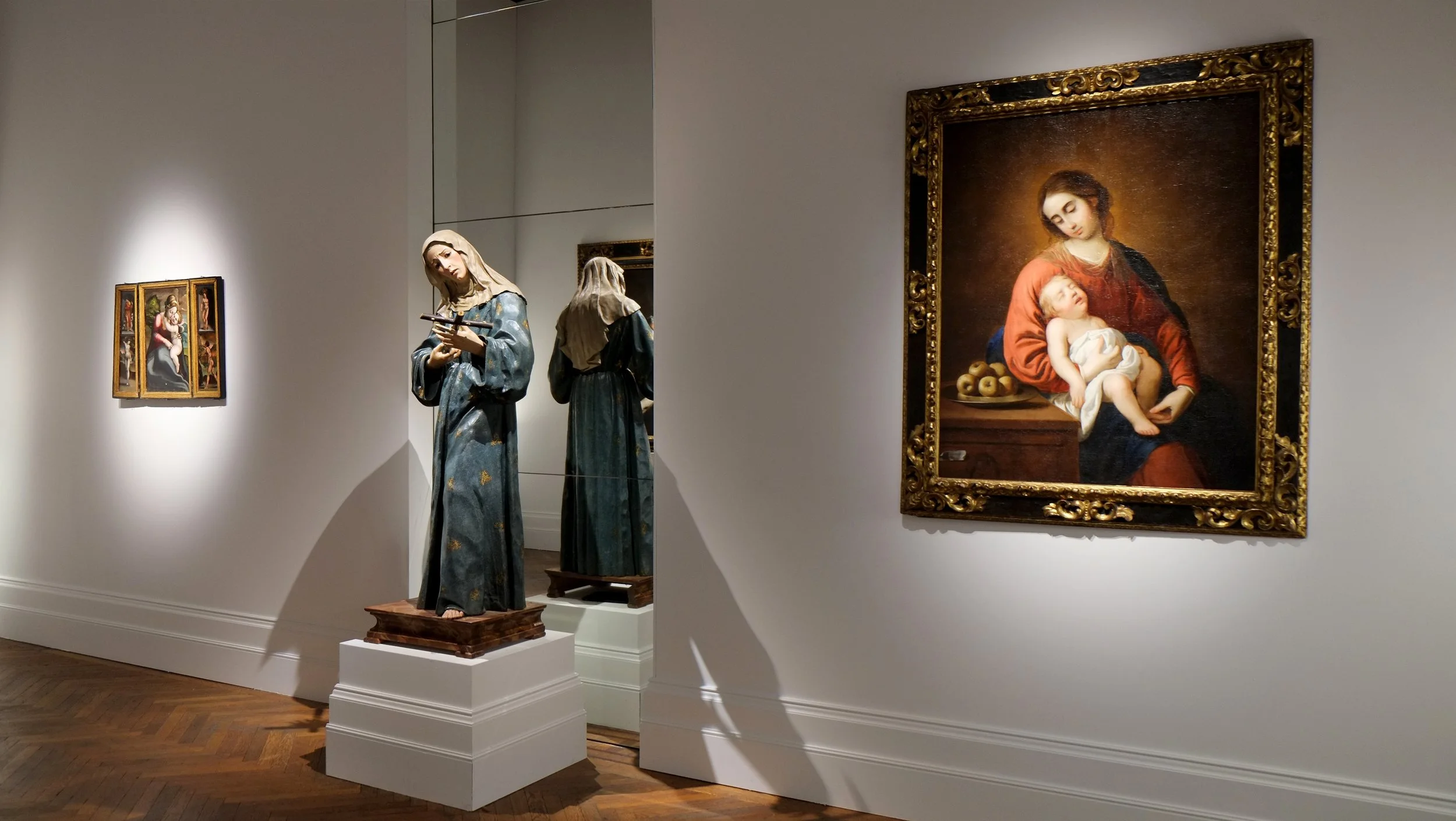 Contemporary gallery installation within a period room featuring religious paintings and wooden sculptures displayed under spot lighting, with mirrors and herringbone flooring.