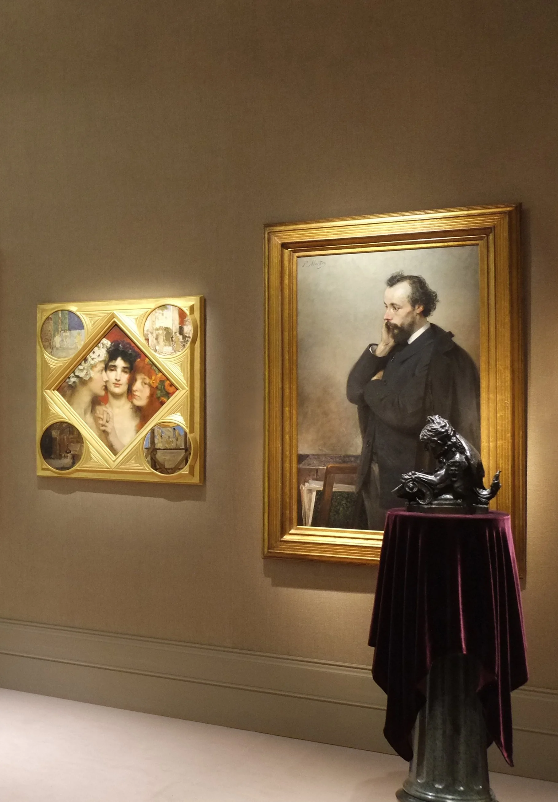 Gallery installation with two paintings and a sculpture displayed on a velvet-draped pedestal against gold fabric walls.