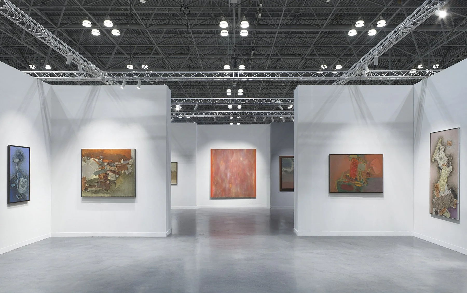 Art fair booth at The Armory Show New York with white and grey walls, a polished concrete floor, and Indian Modern and contemporary abstract paintings displayed under truss-mounted spotlights in an industrial hall setting.