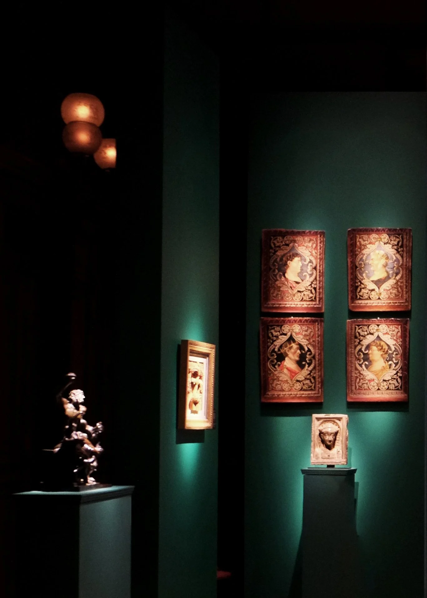 Museum period room with a temporary installation of framed artworks and a sculpture displayed against deep green walls under dramatic lighting.