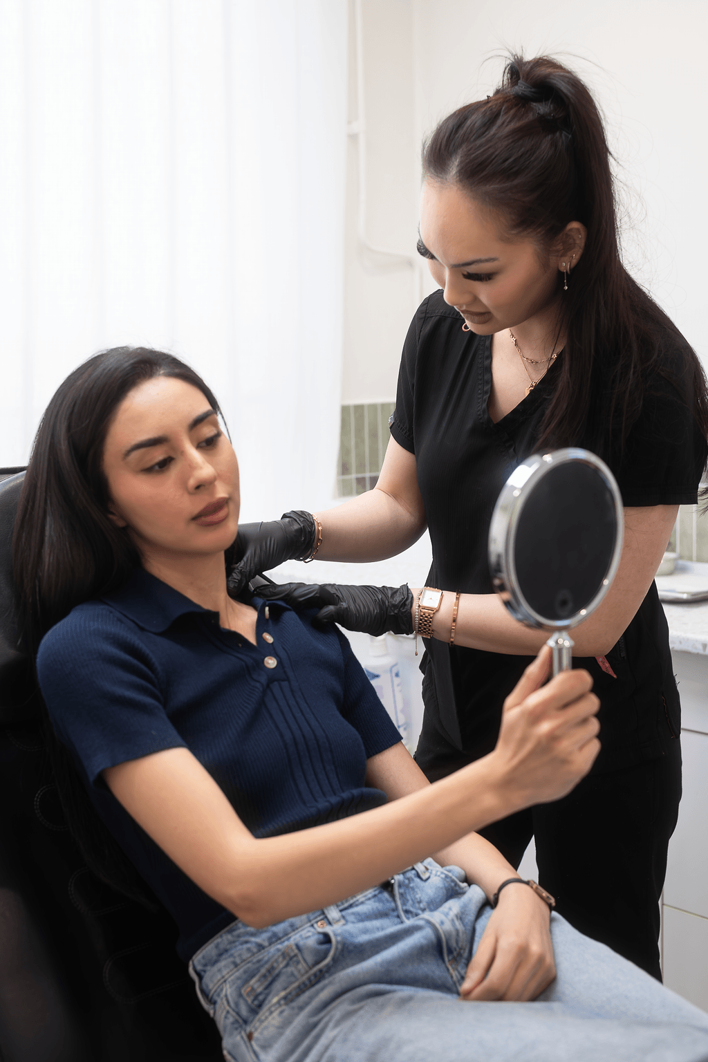 A woman in black scrubs and wearing gloves is examining a woman's neck and shoulders who is sitting in a chair holding a mirror