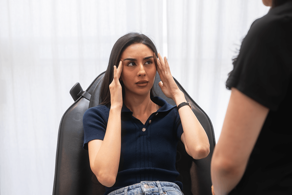 A woman sitting in a chair with a worried expression, holding her head, talking to another person.