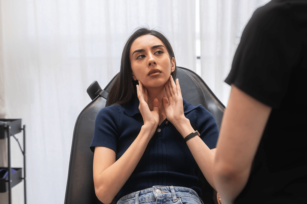 A young woman with dark hair, sitting in a chair, touching her jaw with an expression of concern or discomfort