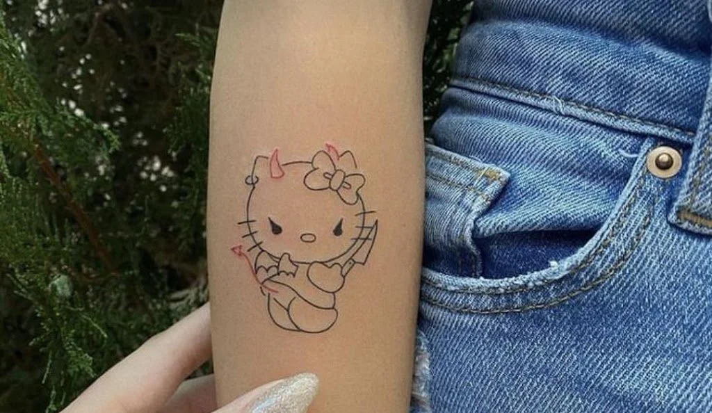 Tattoo of Hello Kitty with devil horns and a bow on her head, holding a bow and arrow, on a person's forearm.